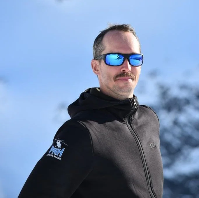 Man wearing reflective sunglasses and a black jacket outdoors in a snowy, mountainous area.