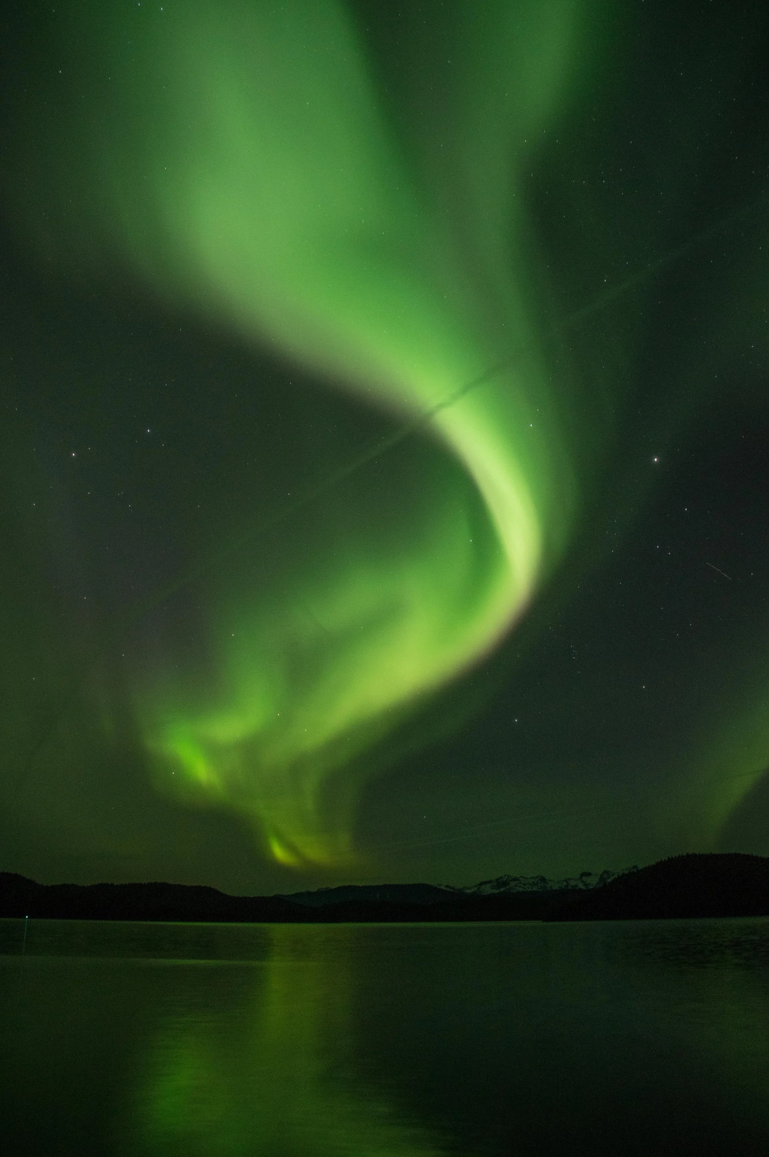 Northern lights (aurora borealis) in Cordova, Alaska glowing green in the night sky above a body of water with mountains in the distance.