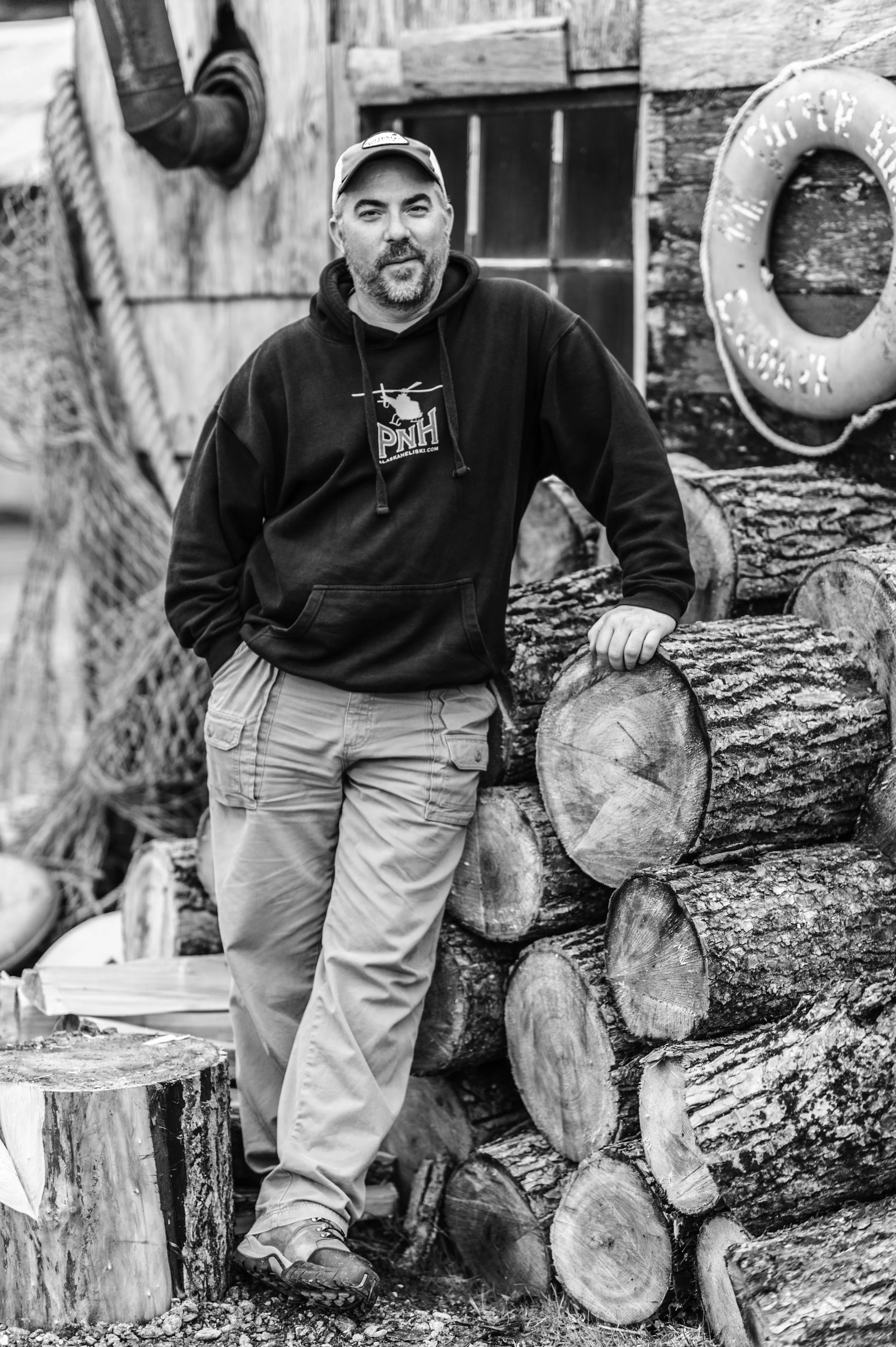 A man leans against a pile of logs, wearing a hoodie and cap, in an outdoor setting with a rustic wooden building and a life preserver in the background.
