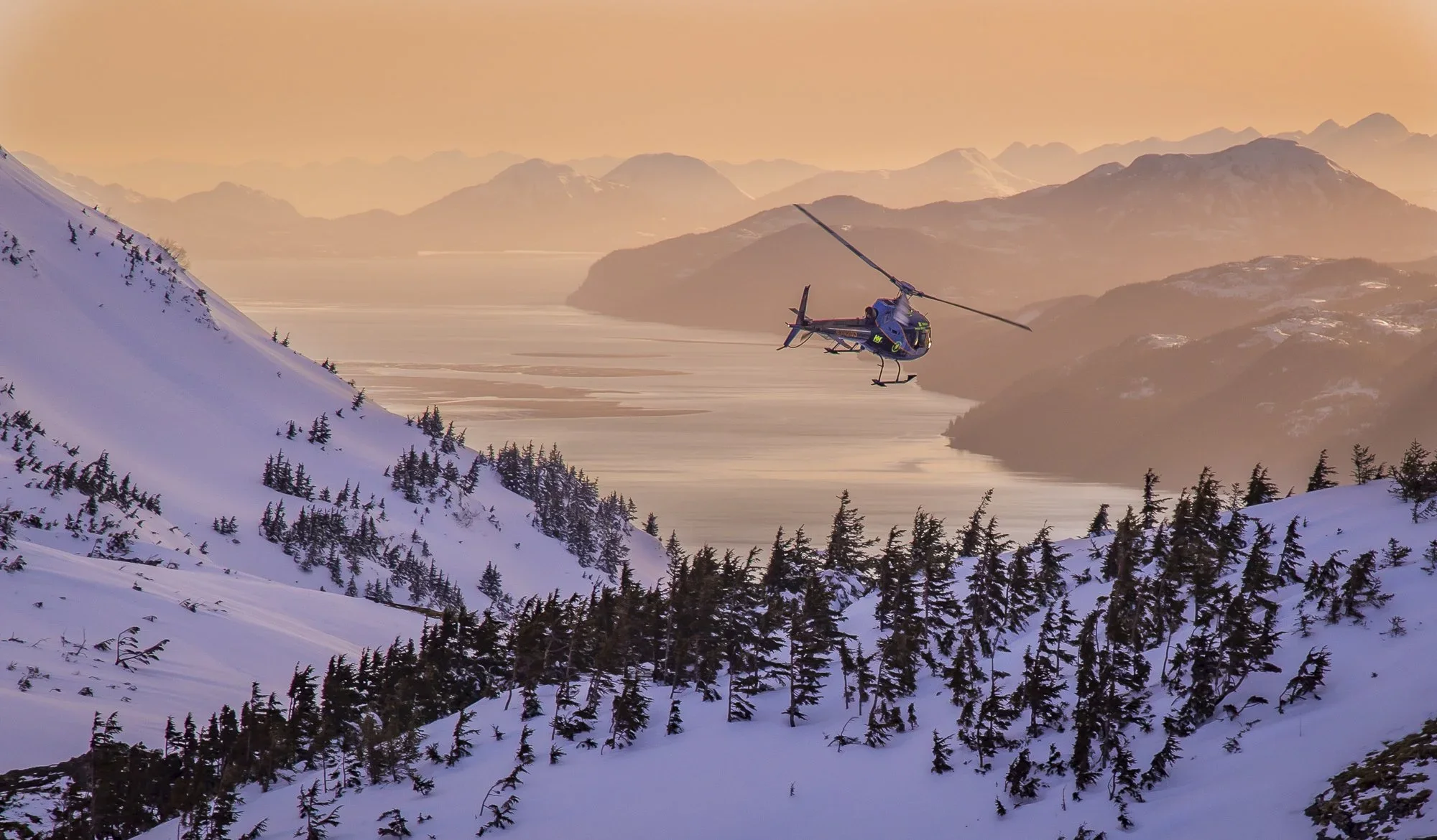 A Points North Helicopter flying over snowy mountain landscape in Alaska at sunset with distant mountains and water bodies.