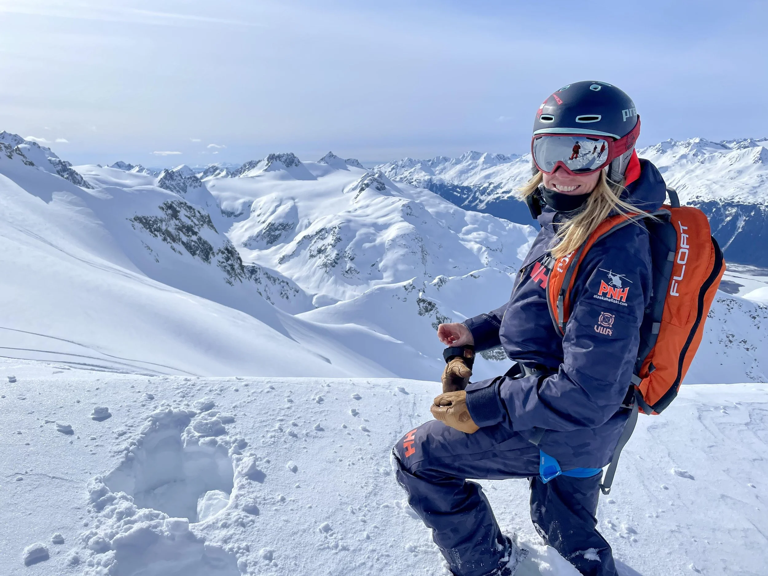A PNH guide in winter gear and ski helmet kneels on snow-covered mountain terrain with snow-capped peaks in the background.