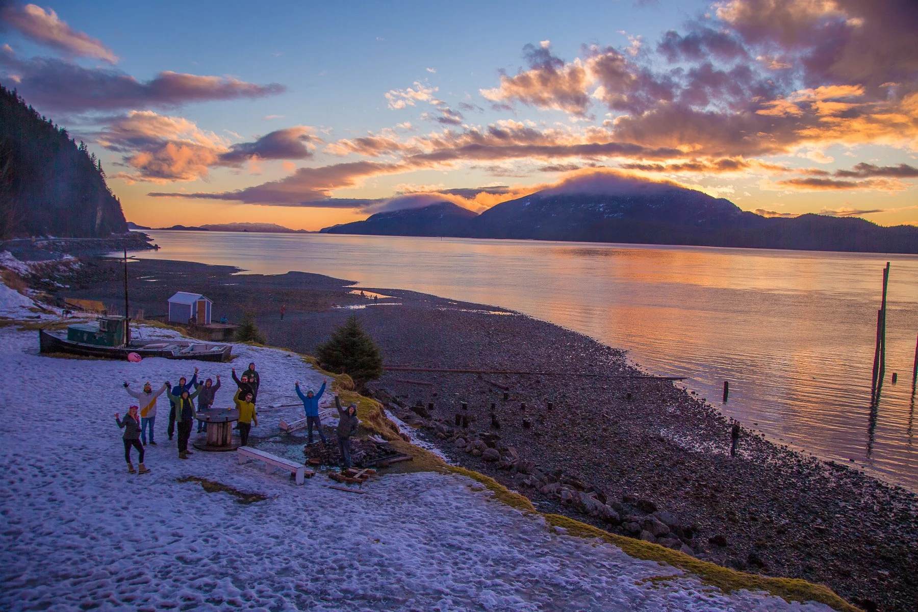 Group of people celebrating on snow-covered beach at sunset with mountains and calm water in the background.