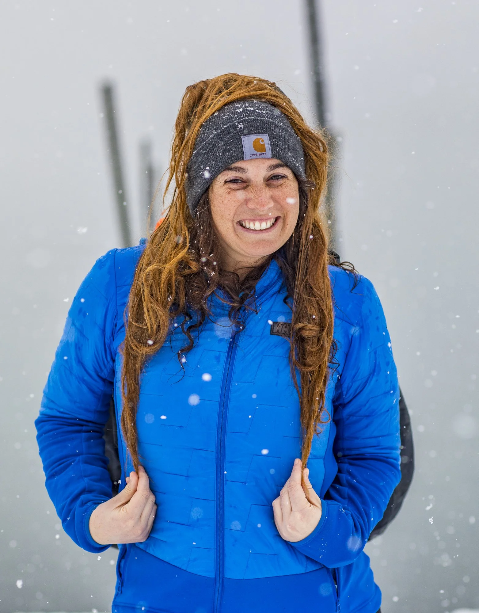 A smiling woman with red hair, wearing a gray Carhartt beanie and a blue jacket, standing in snowy weather.