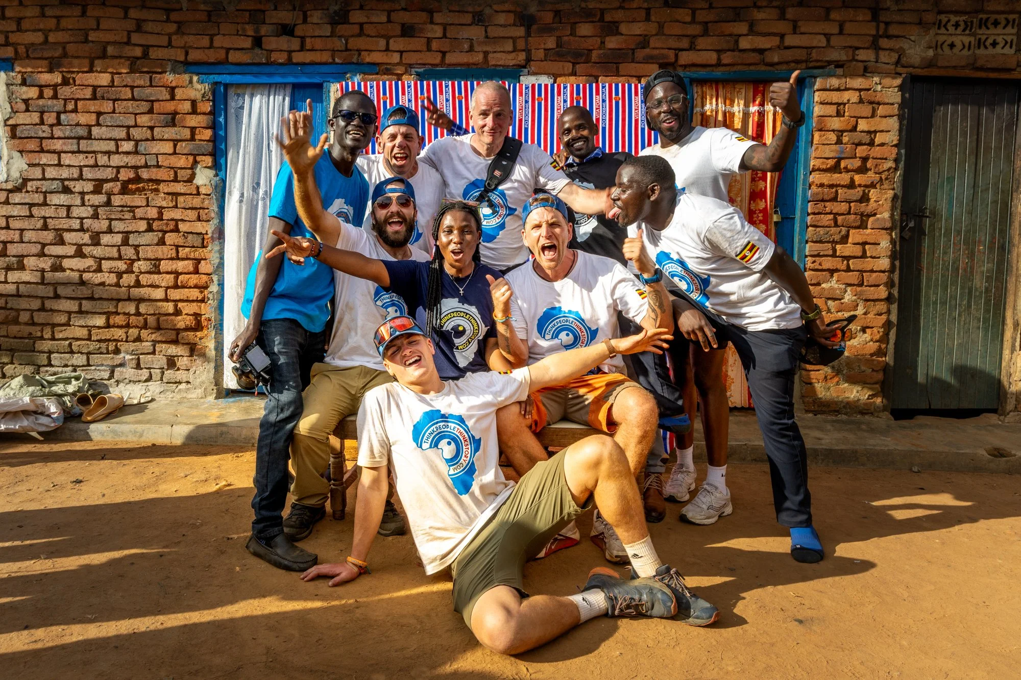 group of diverse people celebrating outdoors in front of a brick building, some wearing matching white T-shirts with a blue logo, smiling, making gestures of excitement and joy.