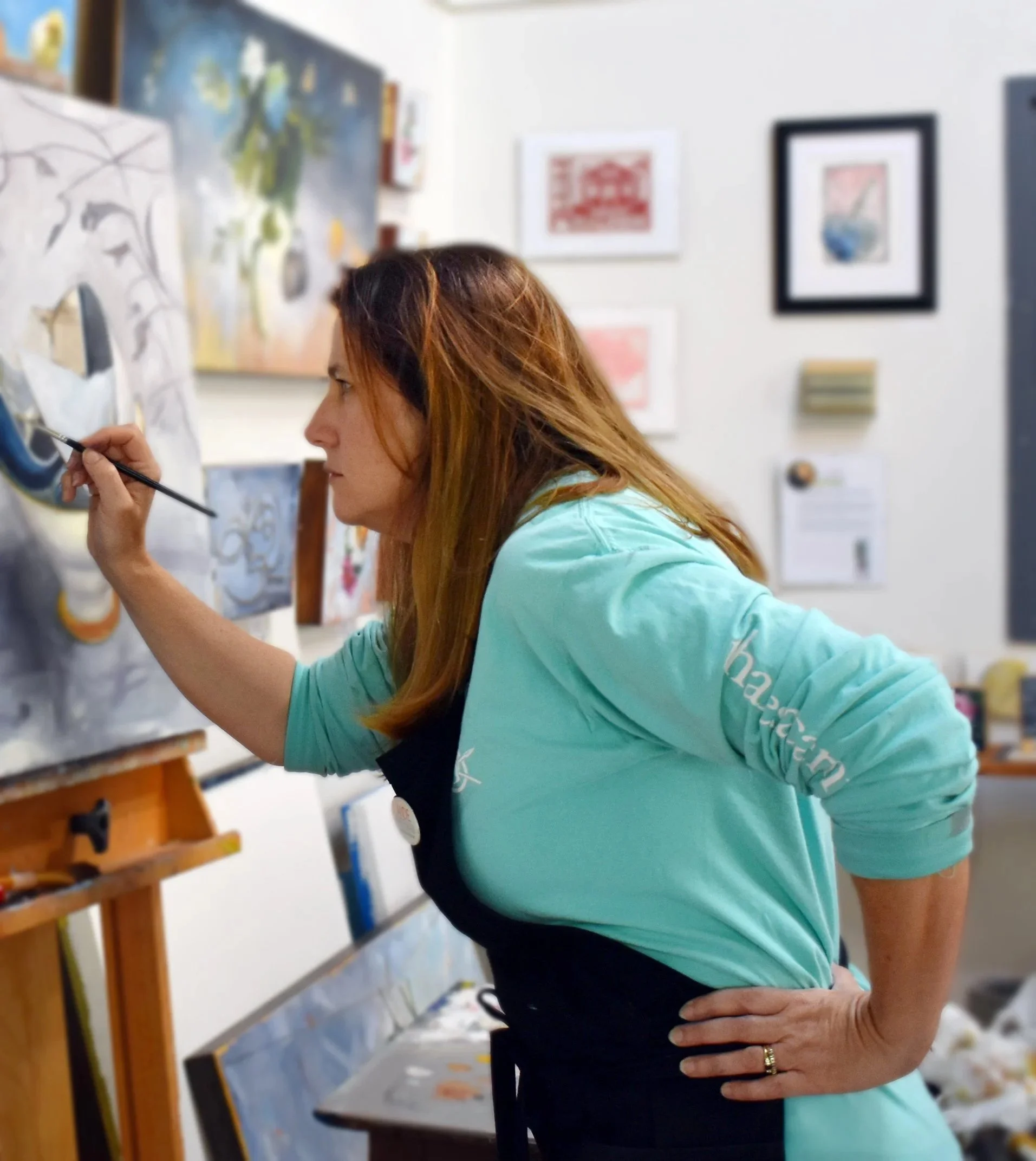 Andie Paradis Freeman painting on an oil painting on canvas at an art studio at Artspace in Raleigh, North Carolina wearing a teal shirt with an art painter's apron.