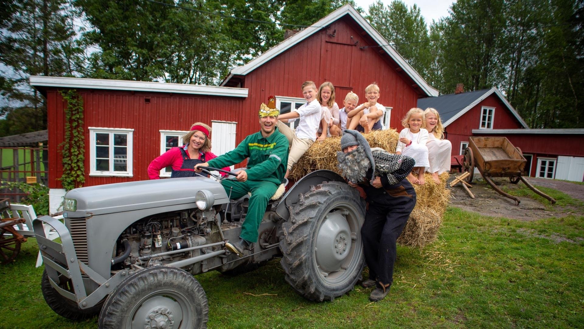 Group of children and adults in costumes riding on a hay wagon behind a tractor in front of a red barn.