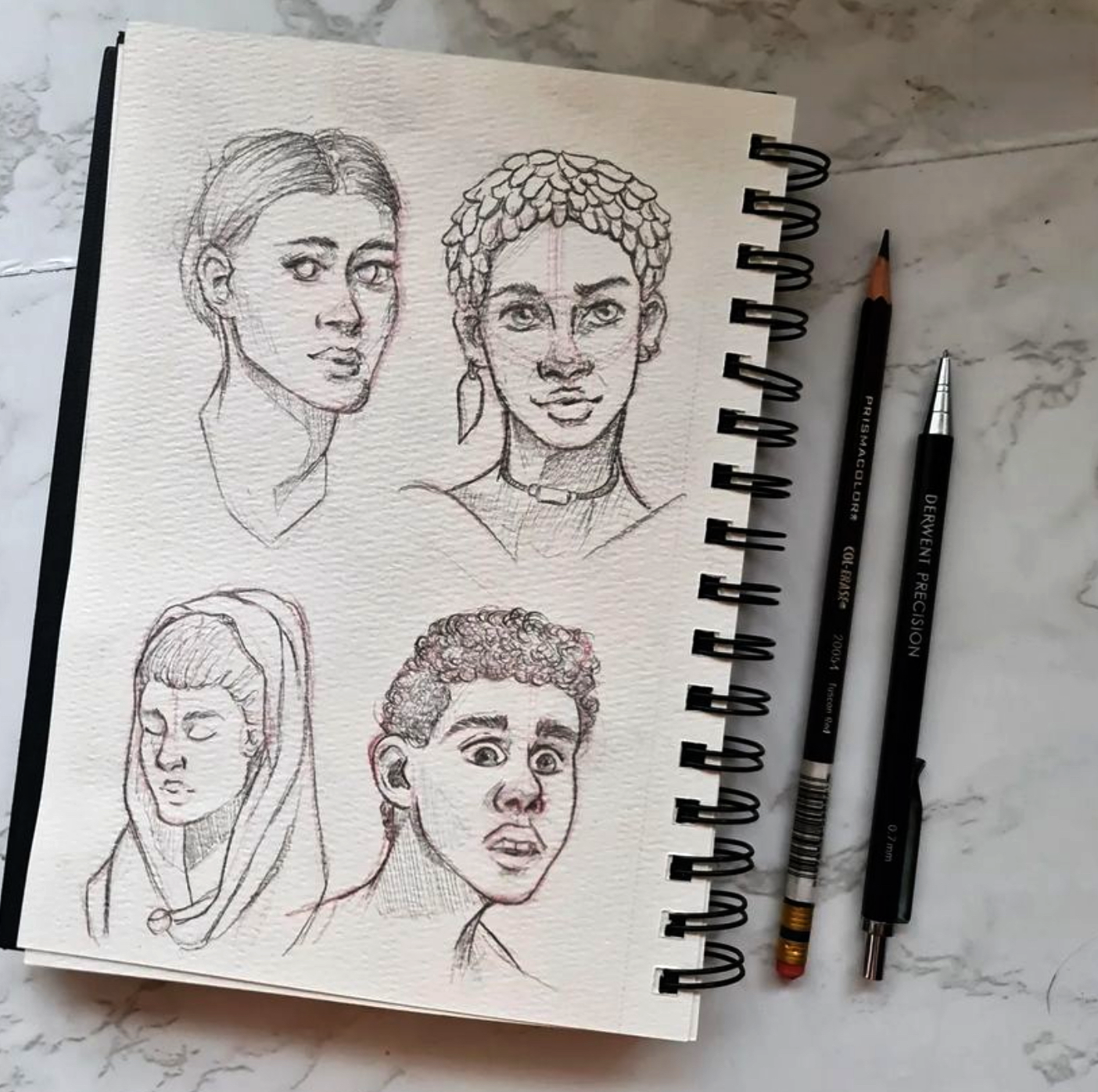 Sketchbook with pencil sketches of four different people's faces, placed on a marble surface with two pencils nearby.