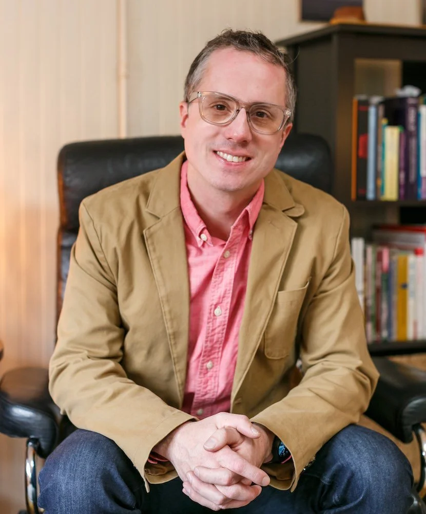 Aaron Good sits in a chair, wearing glasses and a tan blazer over a pink shirt.