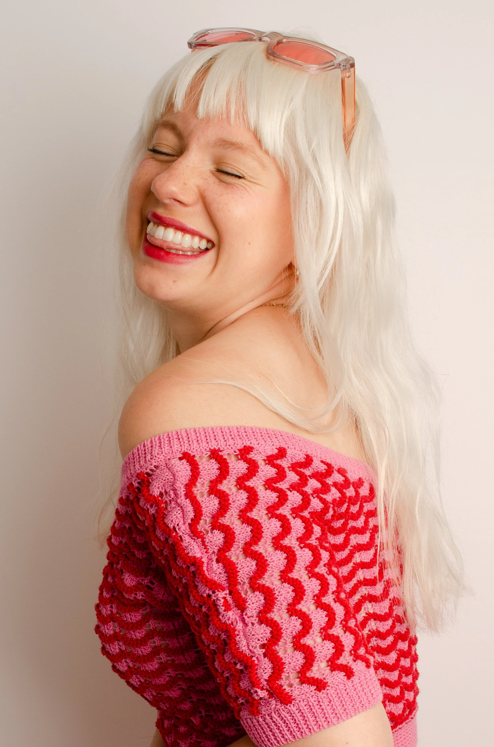 A woman with platinum blonde hair, closed eyes, and a broad smile showing her teeth, appears to be joyful. She is wearing pink sunglasses on her head and a pink knit sweater with red patterns, off her shoulders. The background is plain white.