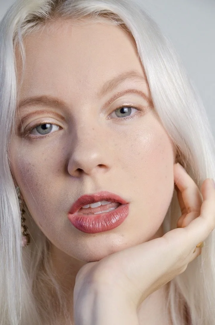Close-up of a young woman with long, platinum blonde hair, fair skin, and blue eyes, resting her face on her hand and looking into the camera.