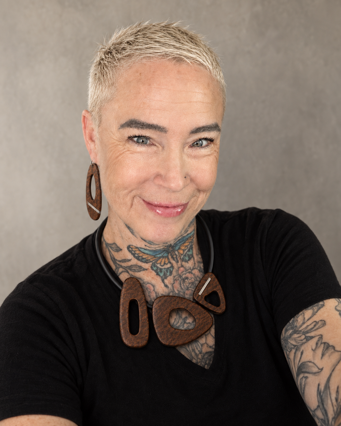 A smiling woman with short blonde hair, tattoos on her neck and arms, wearing large wooden jewelry, a nose ring, and a black shirt, taking a selfie against a plain background.