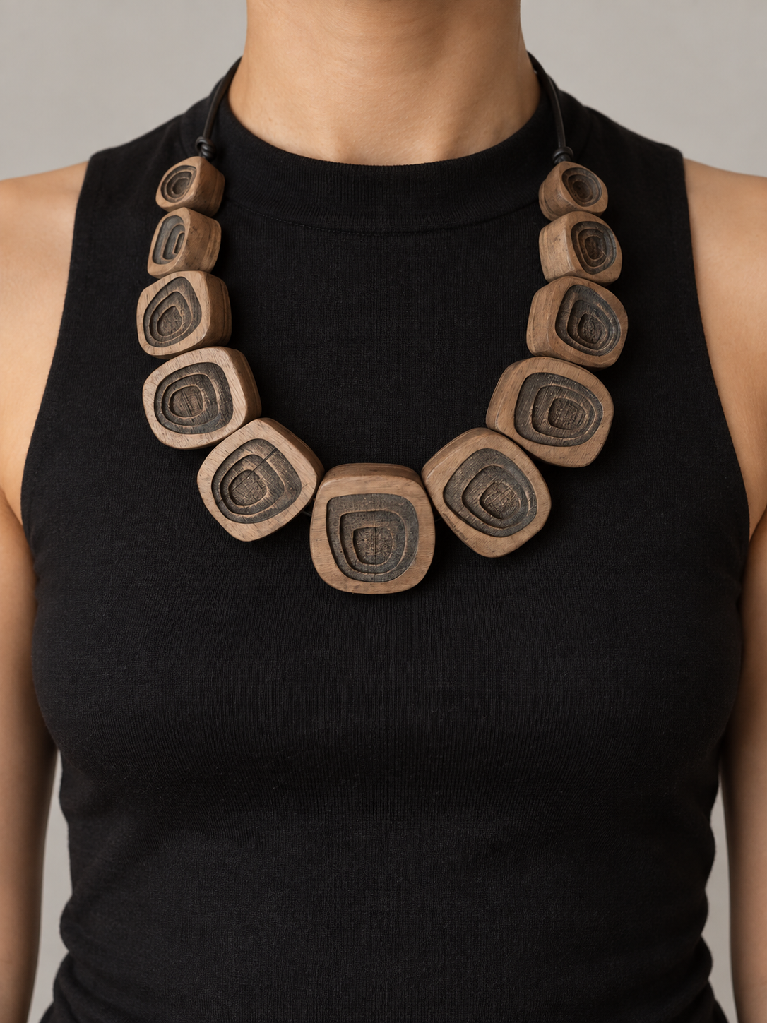 Close-up of a person wearing a black sleeveless top and a large wooden necklace with square beads featuring concentric oval patterns.