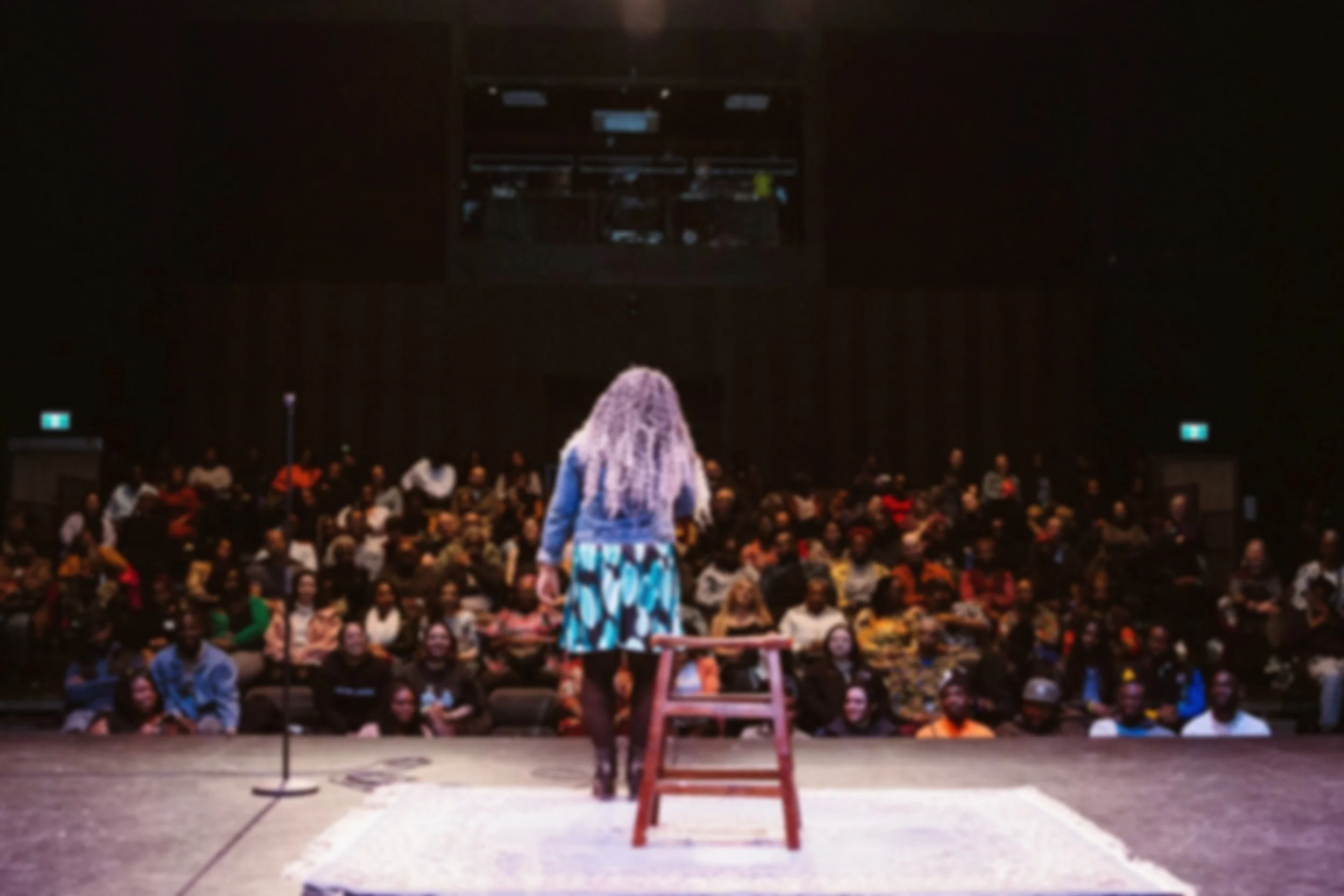 Performer on stage faces a sold-out audience, with her back to the camera.
