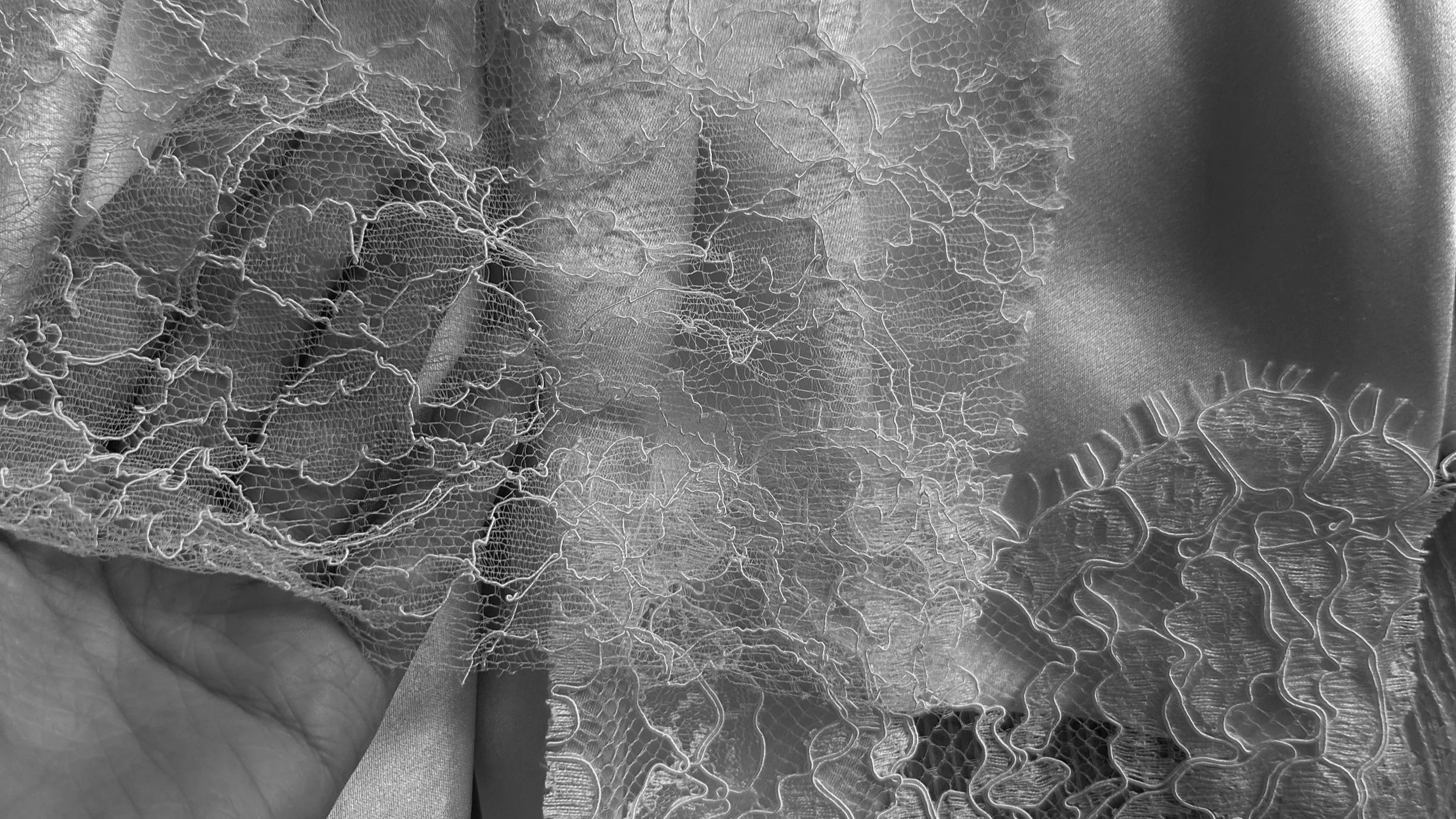 Close-up of a hand holding delicate white lace fabric over a satin background.