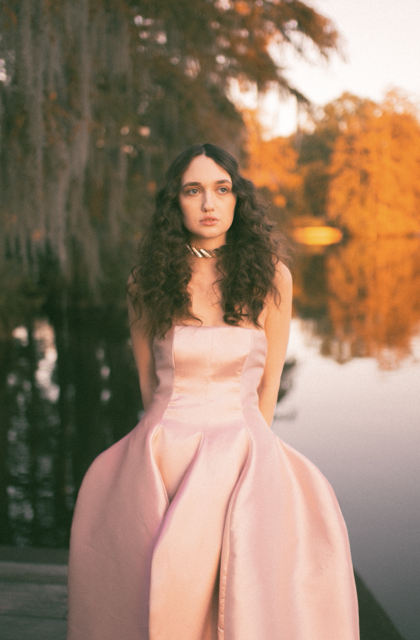 Woman in pink dress standing by a lake with autumn trees in the background.