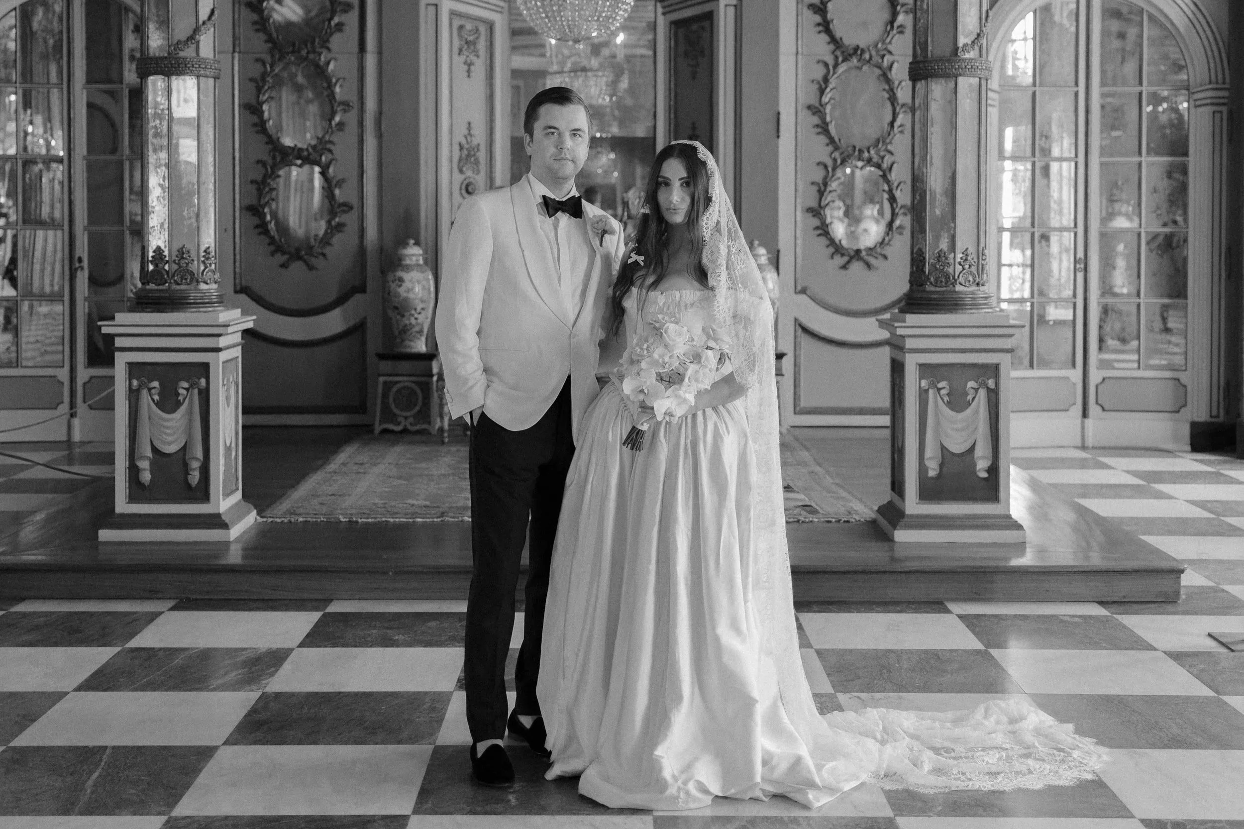 A couple dressed in wedding attire stand in an ornate room with a black and white checkered floor. The man wears a white jacket and black bow tie, while the woman wears a white wedding dress with lace veil, holding a bouquet. The room features decorative mirrors, columns, and chandeliers.