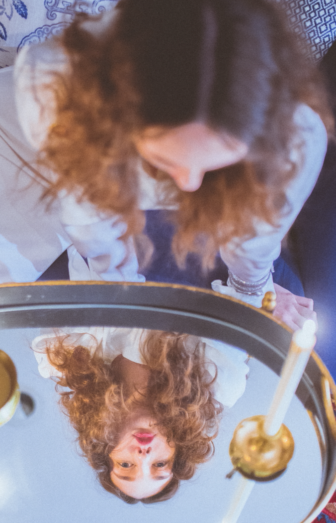 Upside-down reflection of a woman with curly hair in a circular mirror, next to a lit candle.