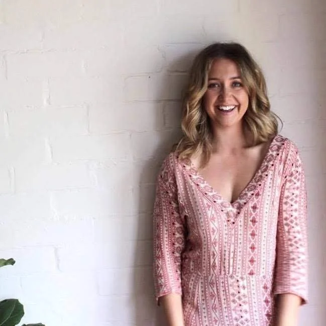 Woman smiling in a patterned dress standing against a white brick wall.