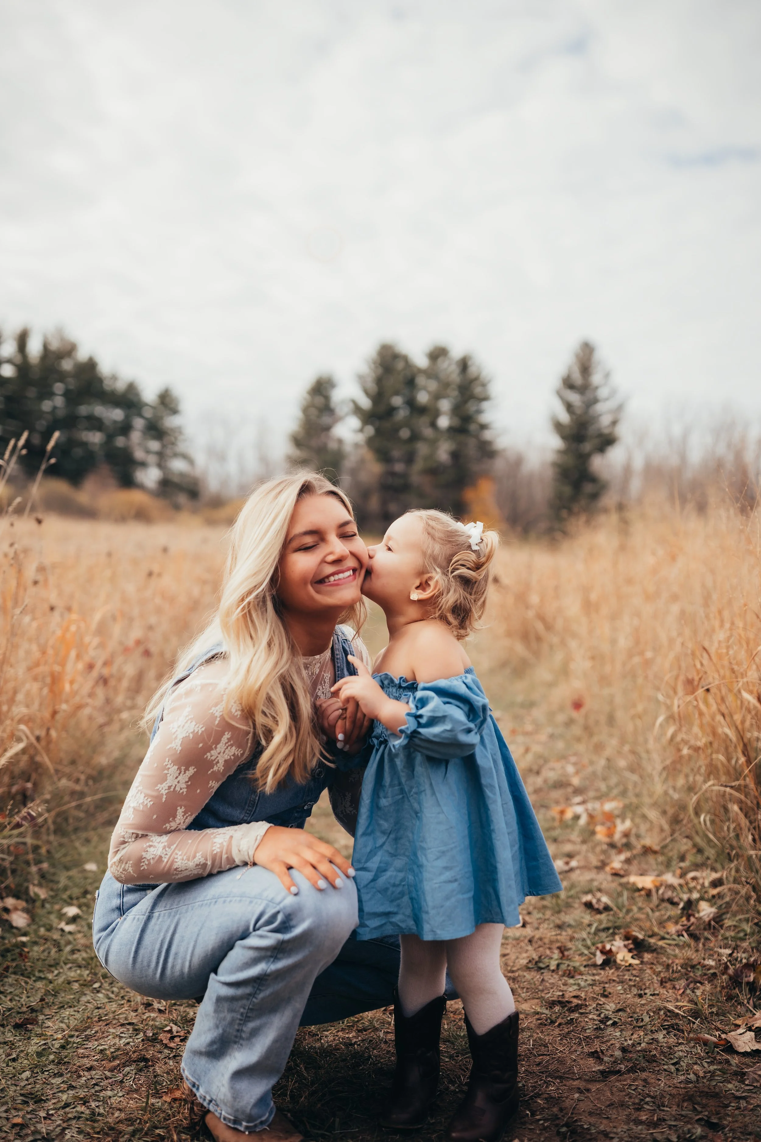 Playful outdoor family photoshoot in a grassy field with mom and young daughter kissing her cheek