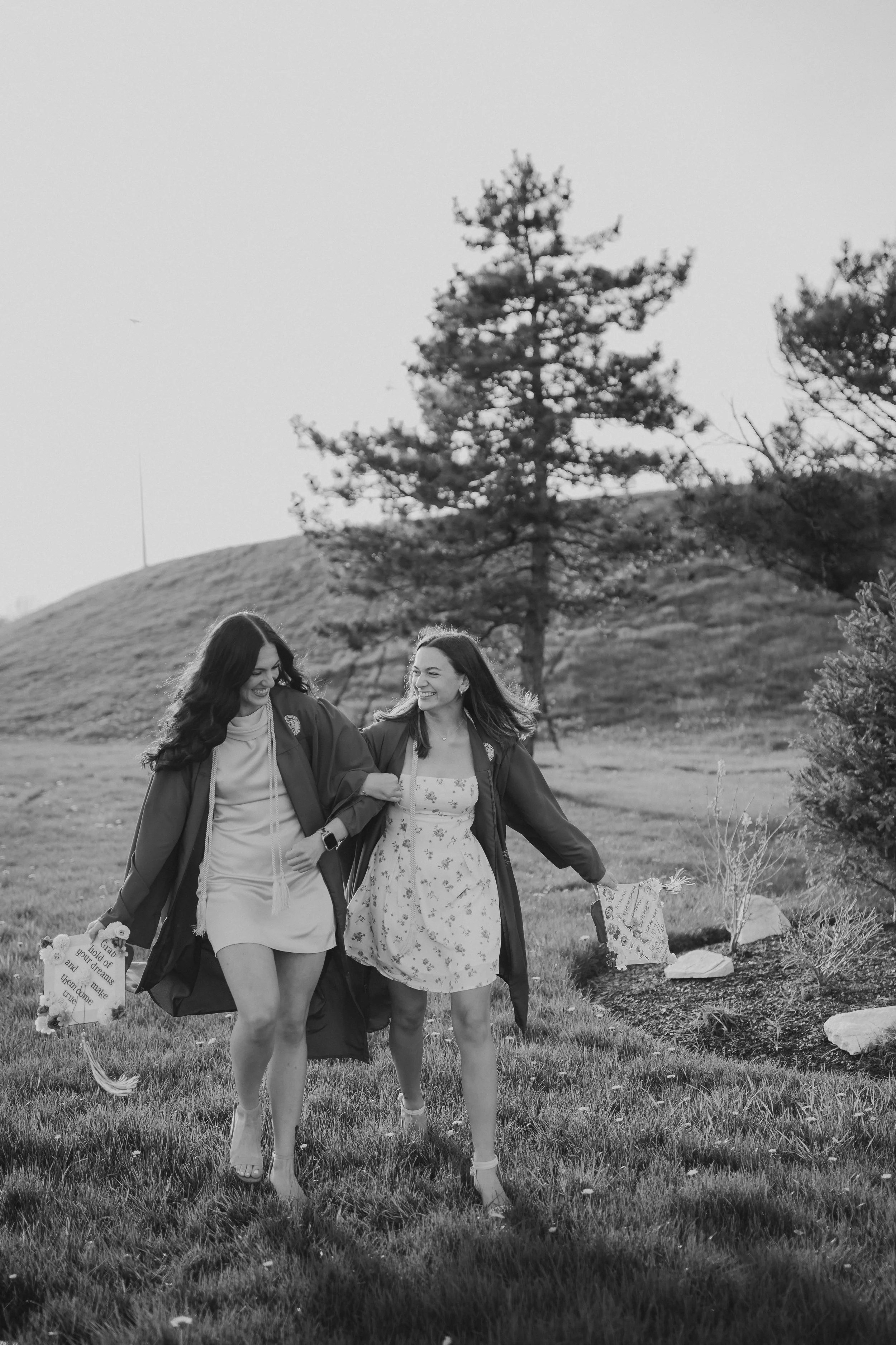 College graduation photoshoot of best friends in their gowns with caps strolling in a field during golden hour