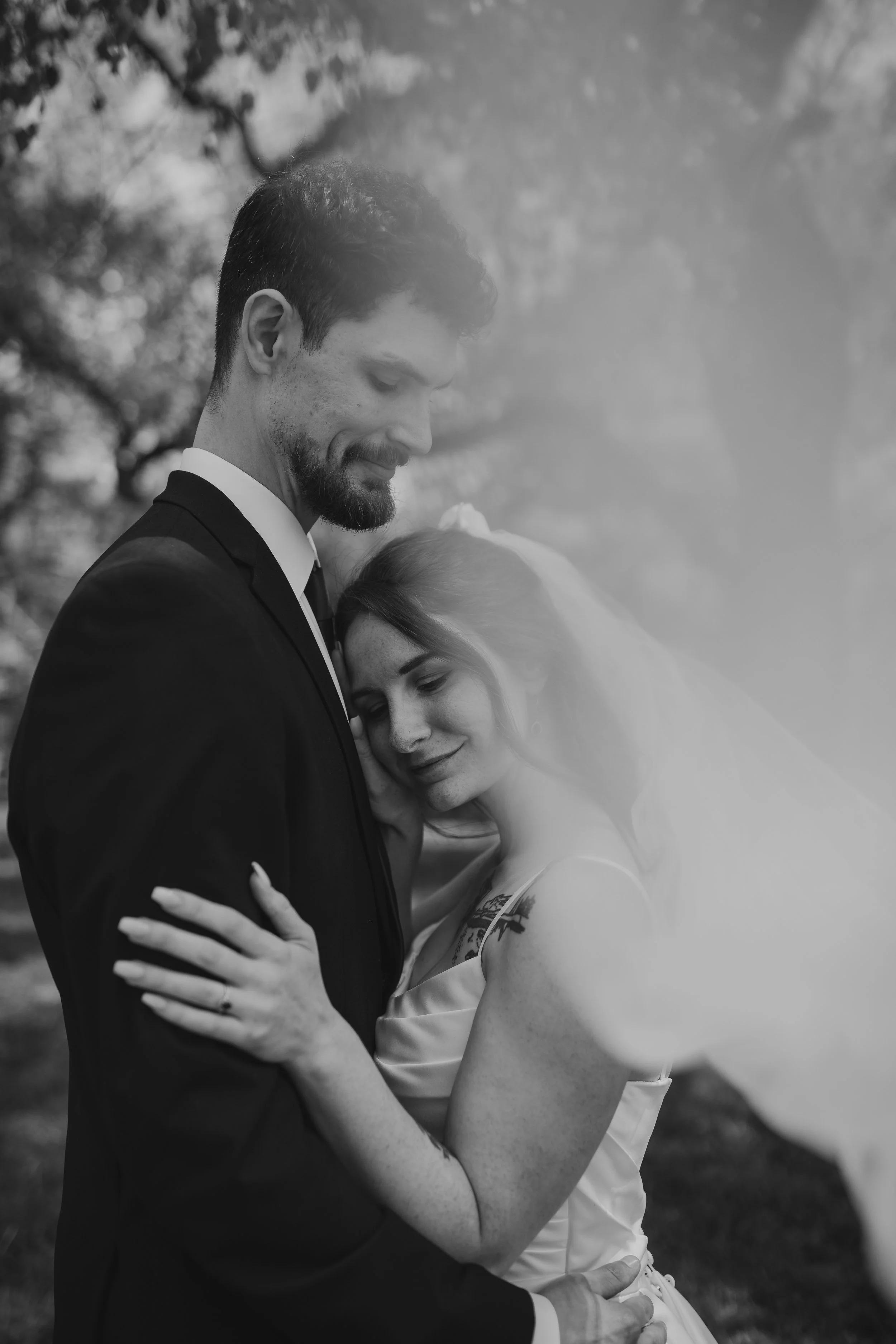 Cinematic black and white photo of bride and groom under the trees wrapped in the bride's veil