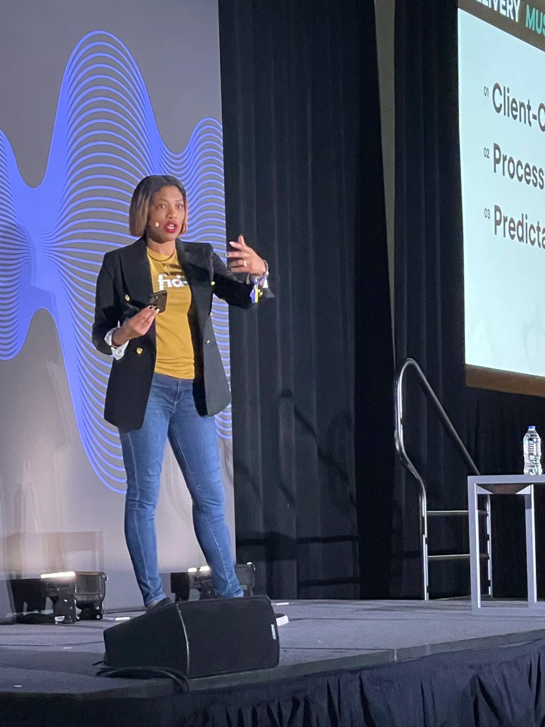 A woman in a black blazer, yellow shirt, and jeans is giving a presentation on stage. She is holding a microphone in one hand and gesturing with her other hand. There is a large screen displaying a slide with numbered points on the right side of the stage.