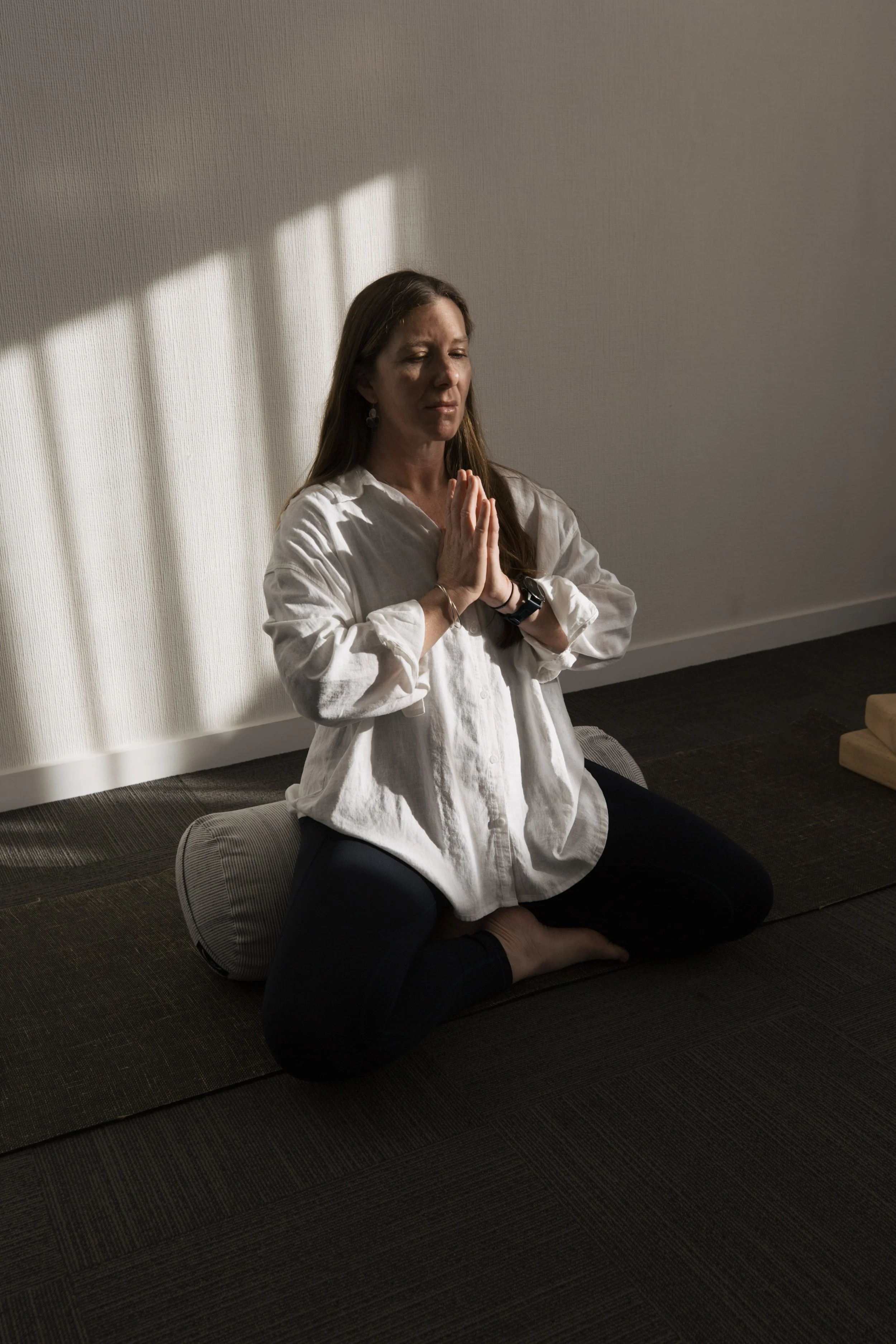Yoga therapist Hayley seated on a bolster with hands in prayer, welcoming contact and connection