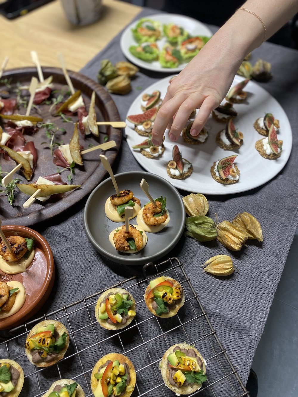 Assorted appetizers on plates and a cooling rack, including mini toasts with toppings, shrimp skewers, and fig slices, arranged on a table for a gathering.