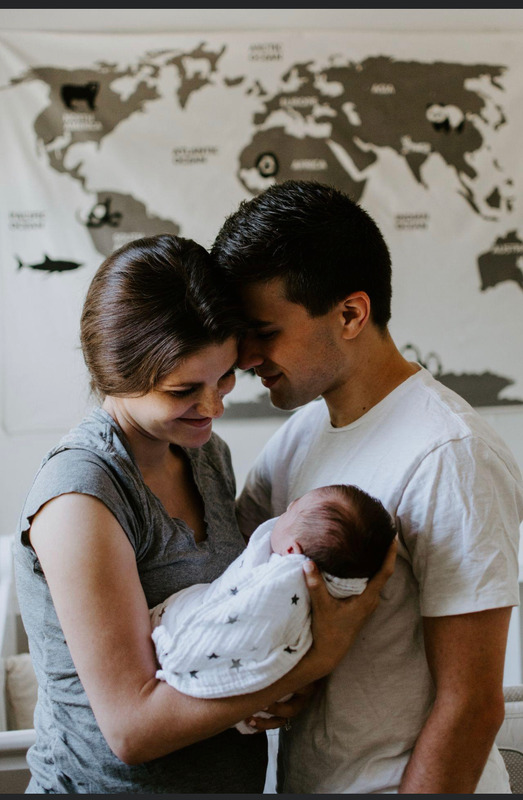 A family of three holding a newborn baby indoors, with a world map on the wall behind them