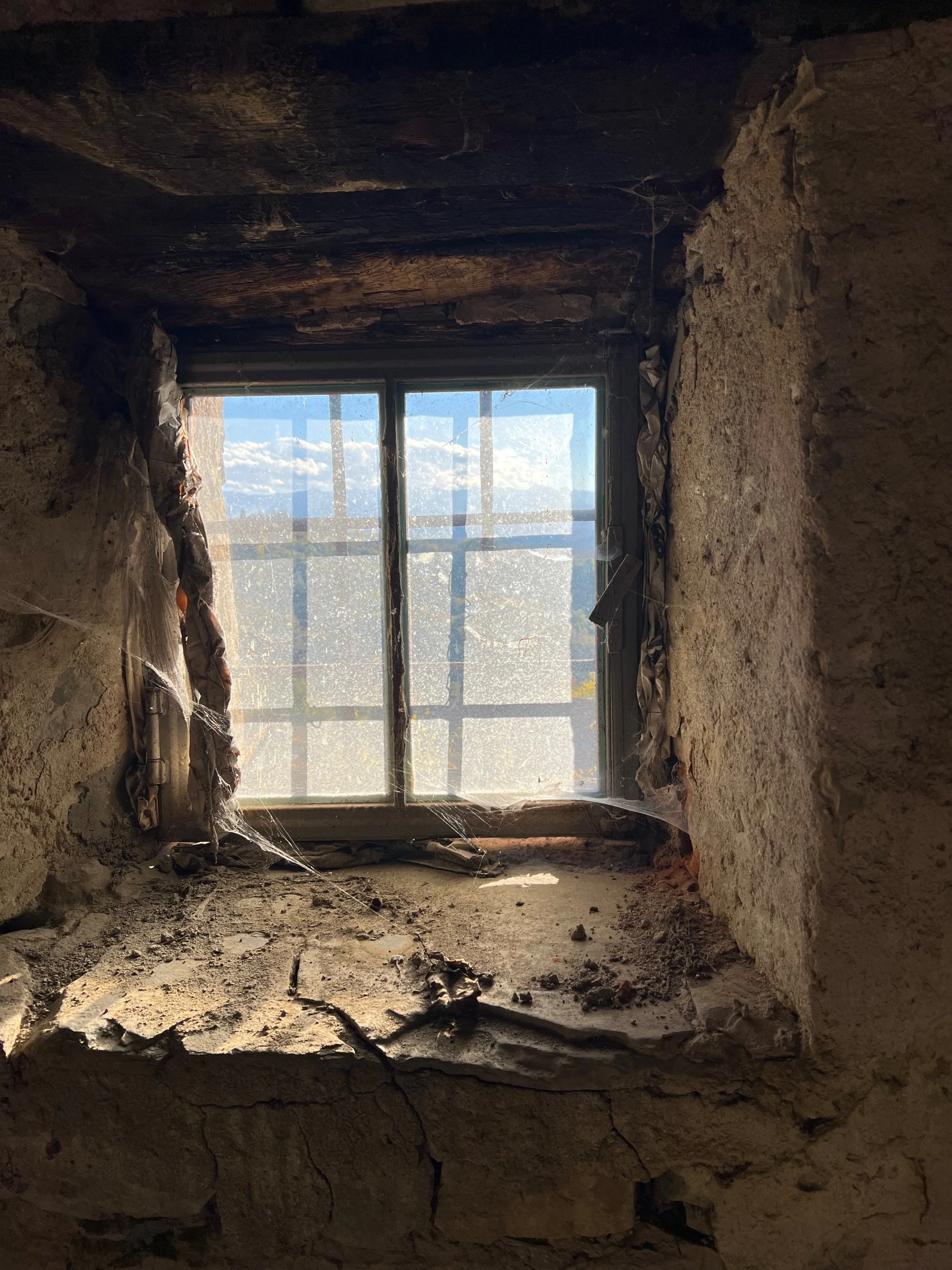 Old, dusty window with a view of blue sky and clouds outside, framed by rough, weathered stone walls and cobwebs.