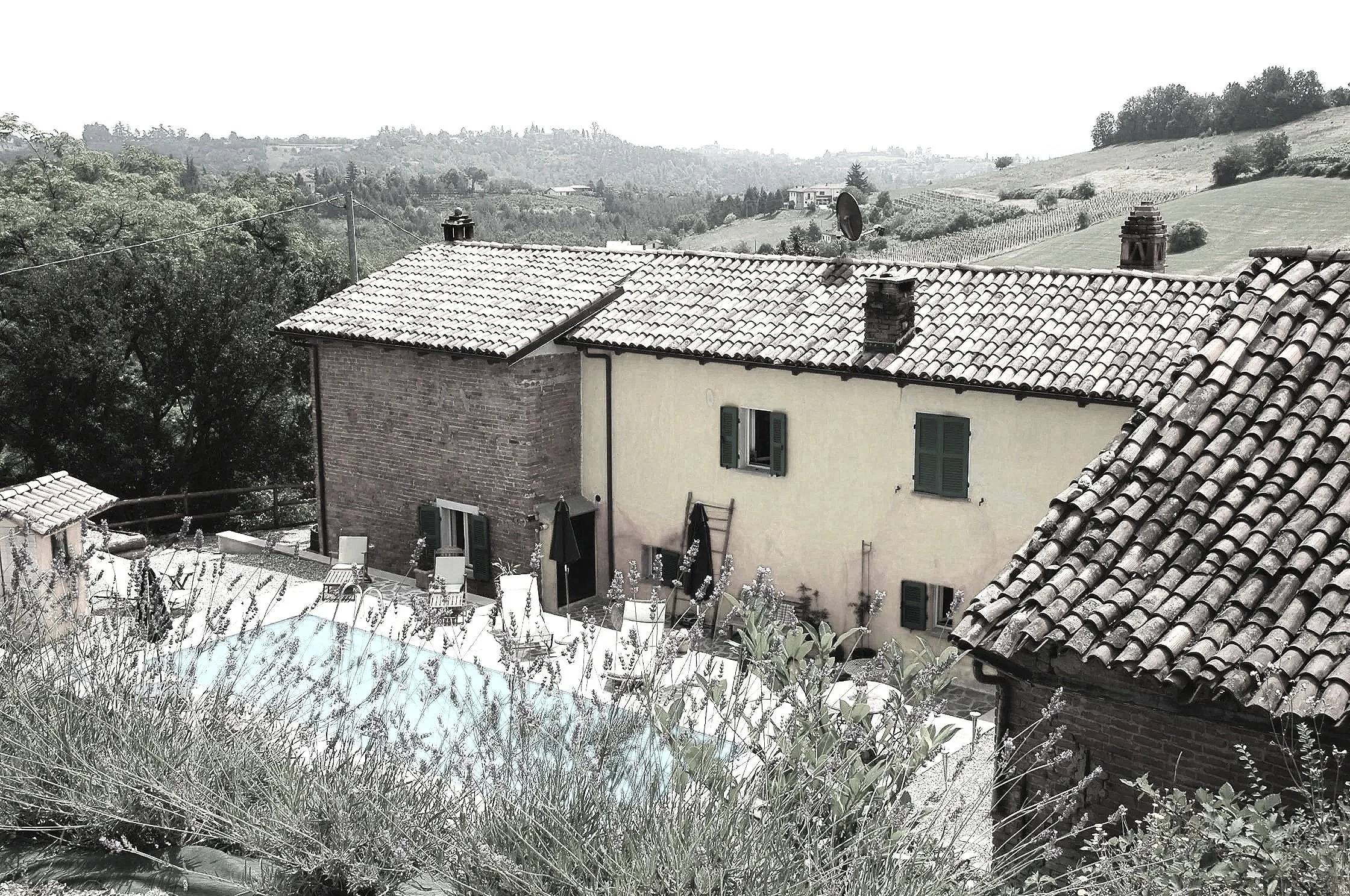 A house with green window shutters, a tiled roof, and a small backyard with outdoor chairs and umbrellas, overlooking rolling hills in a rural area.