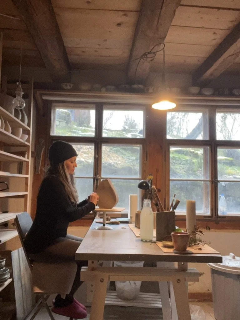 Woman sitting at a wooden table in a rustic pottery studio, working on a piece of pottery, with pottery and tools on the table and shelves behind her.