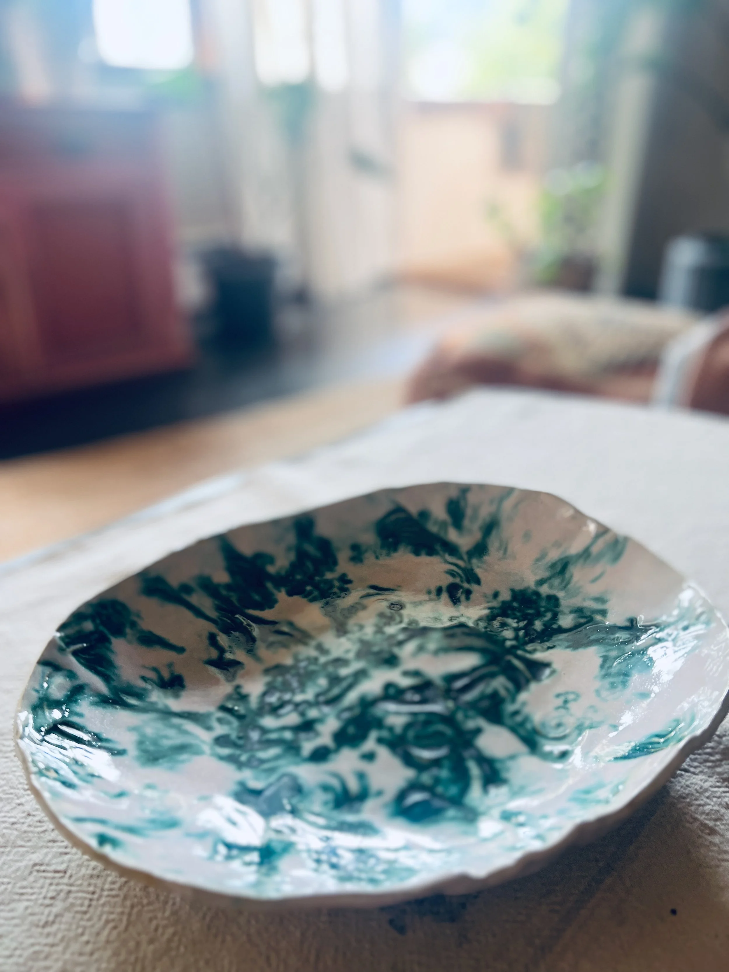 Close-up of a decorative ceramic bowl with blue and white marbled pattern sitting on a beige cloth, with a blurry background of a room with furniture and windows.