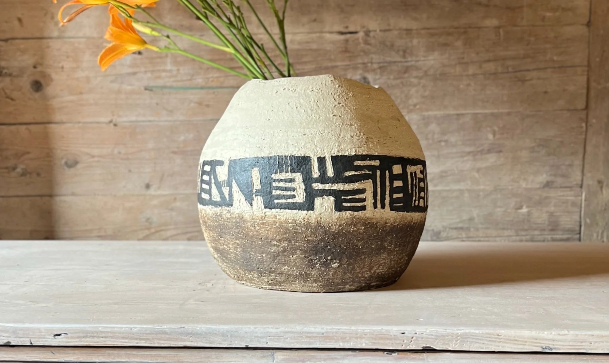 A beige and dark brown ceramic vase with black geometric patterns, holding orange flowers, on a wooden surface against a wooden background.