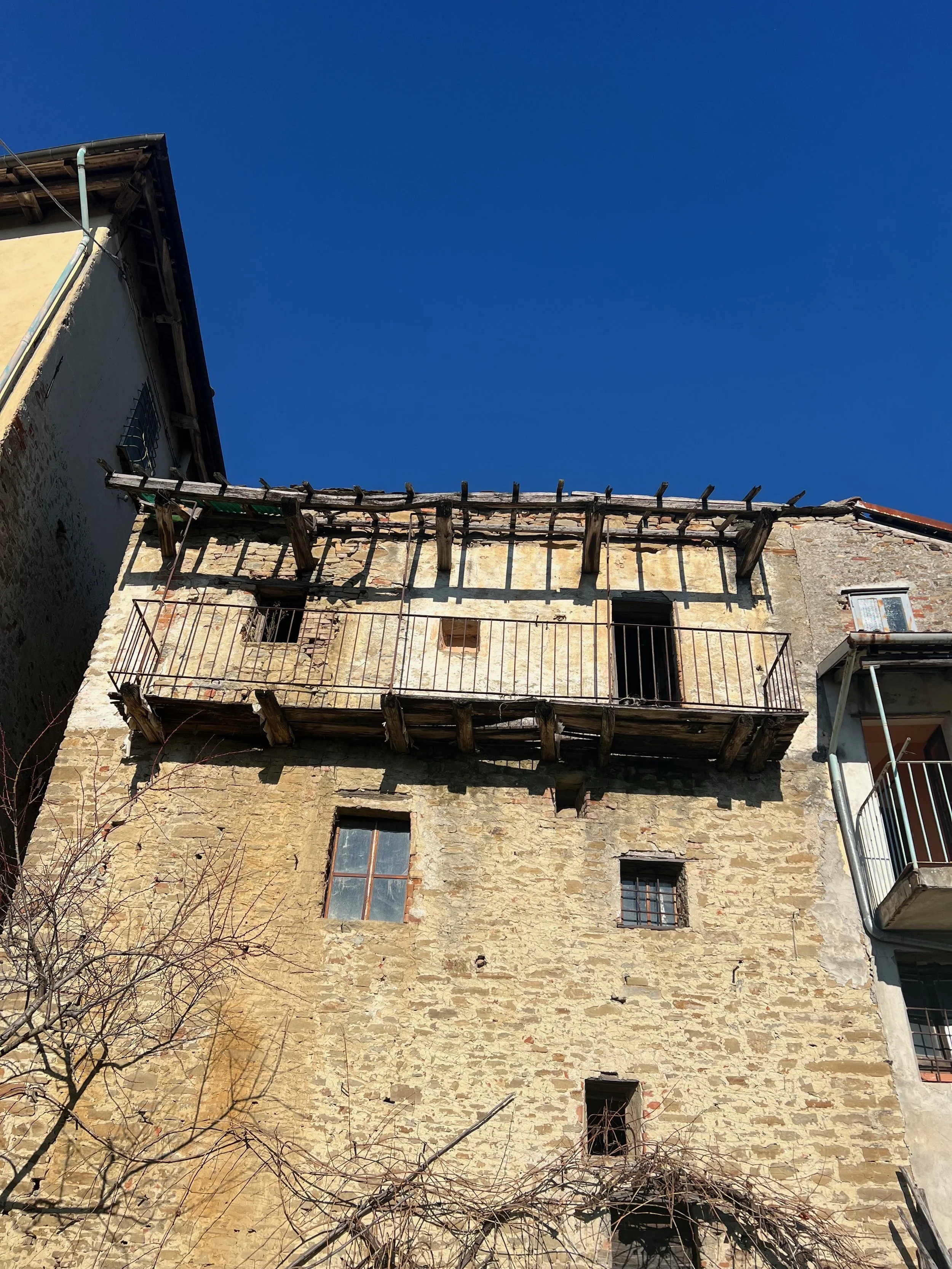 Old stone building with small windows and wooden balcony with railing, under a clear blue sky.