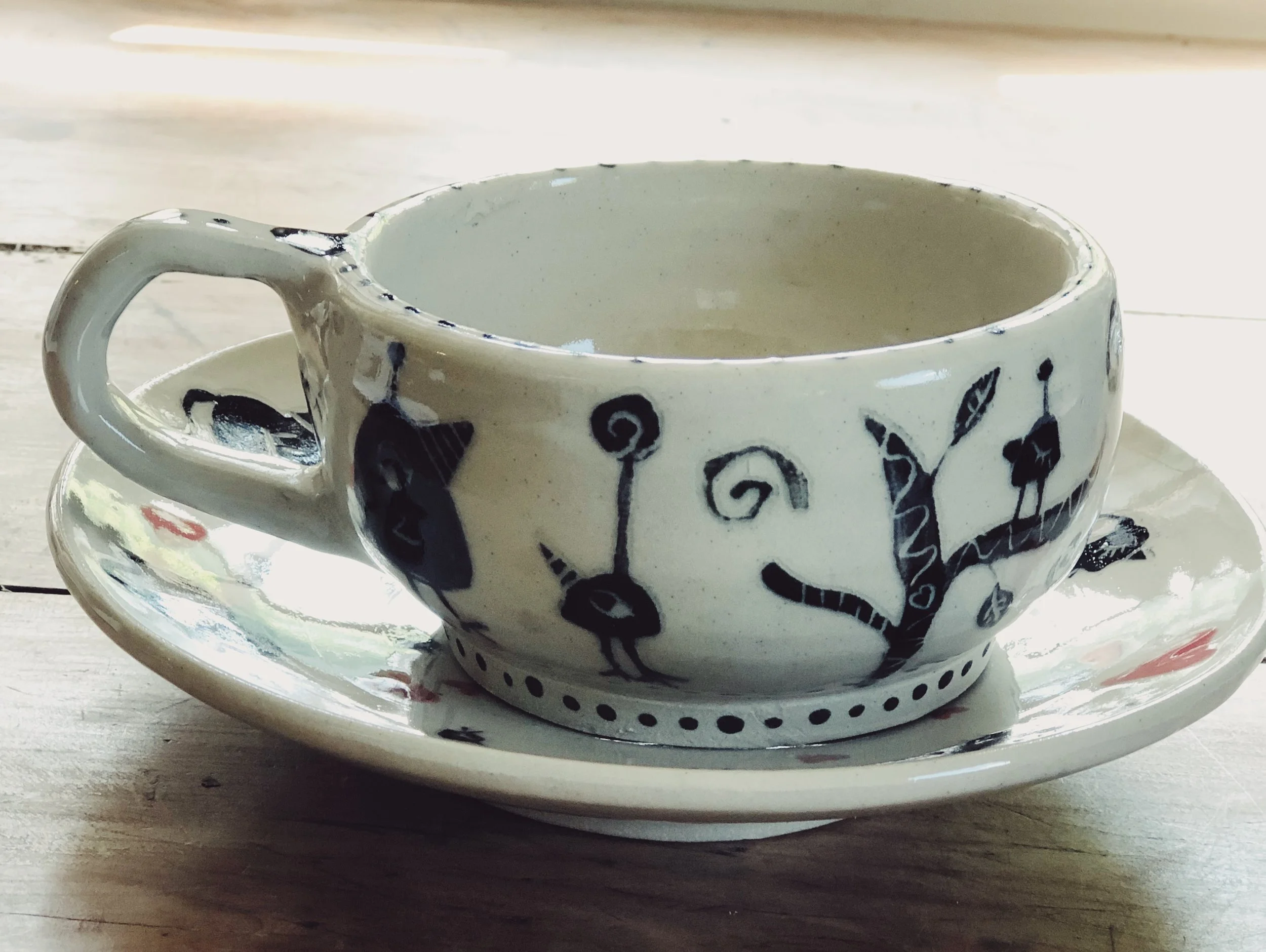 A ceramic cup with a white background and black abstract animal and plant designs, placed on a matching saucer on a wooden surface.