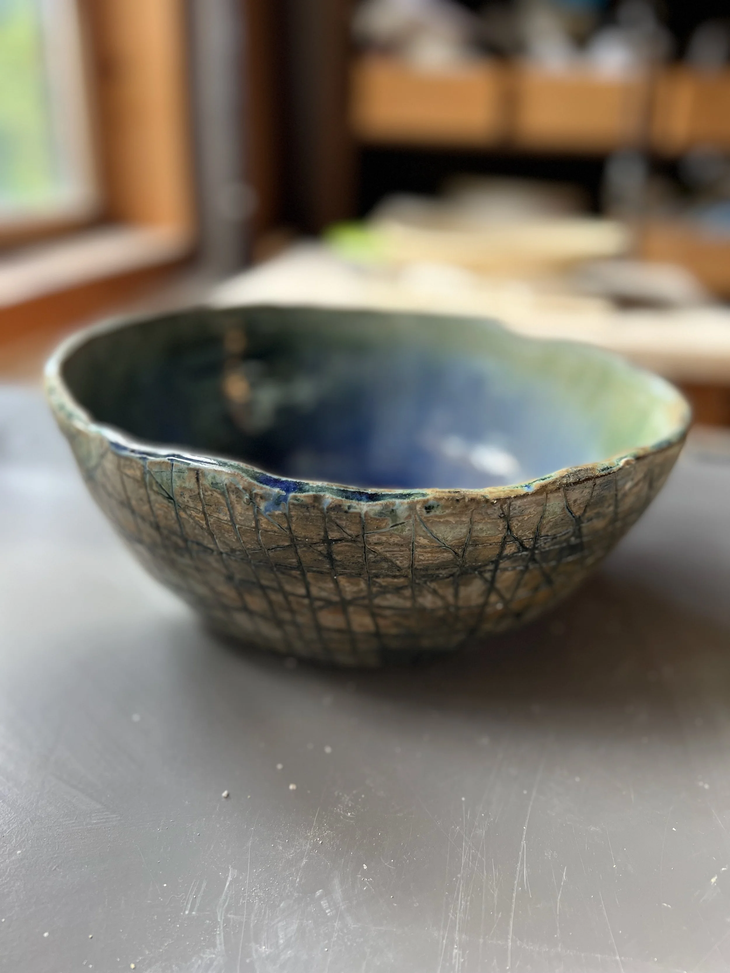 A handcrafted ceramic bowl with a textured exterior and a glossy, multicolored interior, placed on a gray surface indoors.