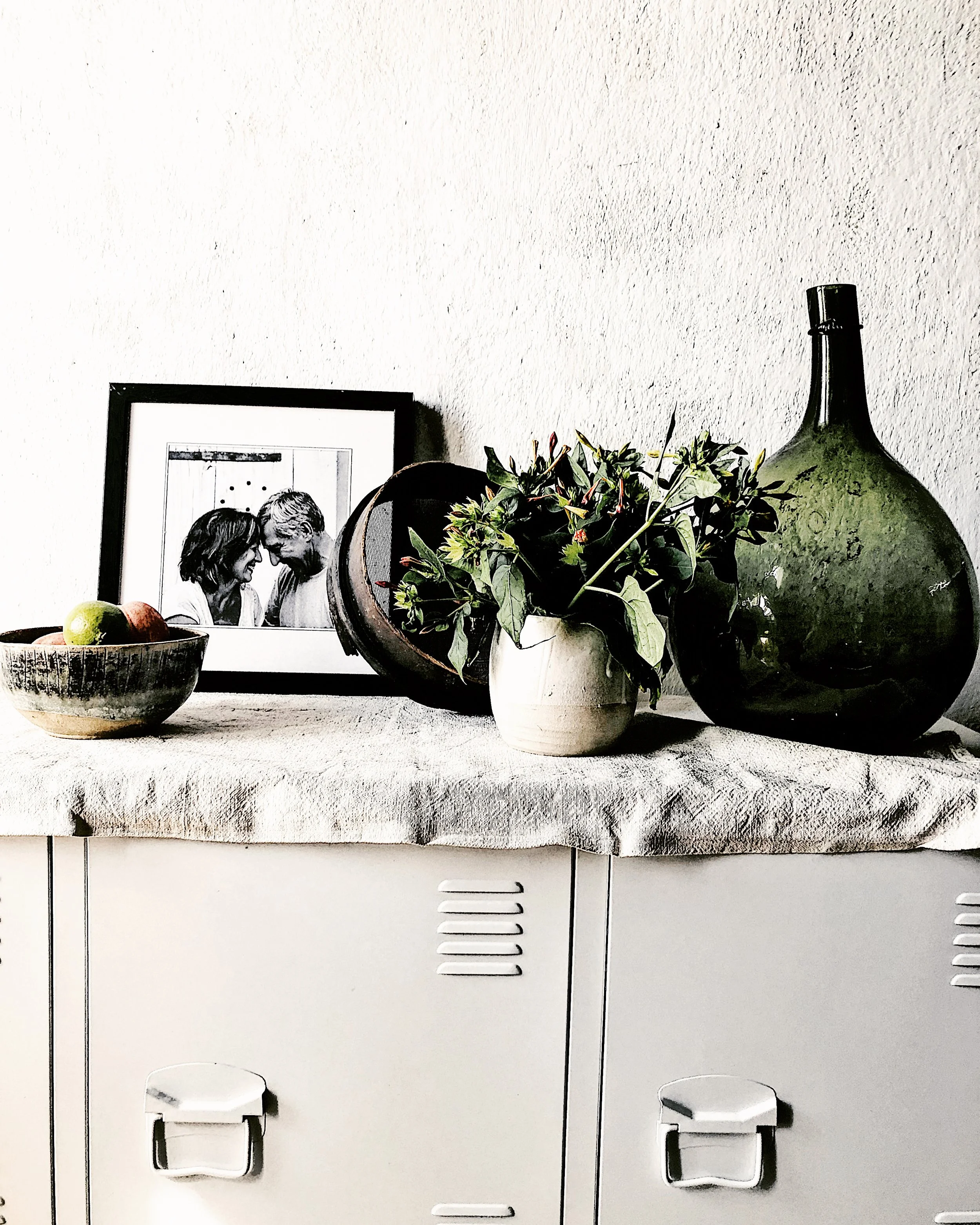 Decorative display on a white surface including a framed black-and-white photo of two people, a small bowl with apples, a white pot with green leafy plant, a large green glass bottle, and a black ceramic pot.