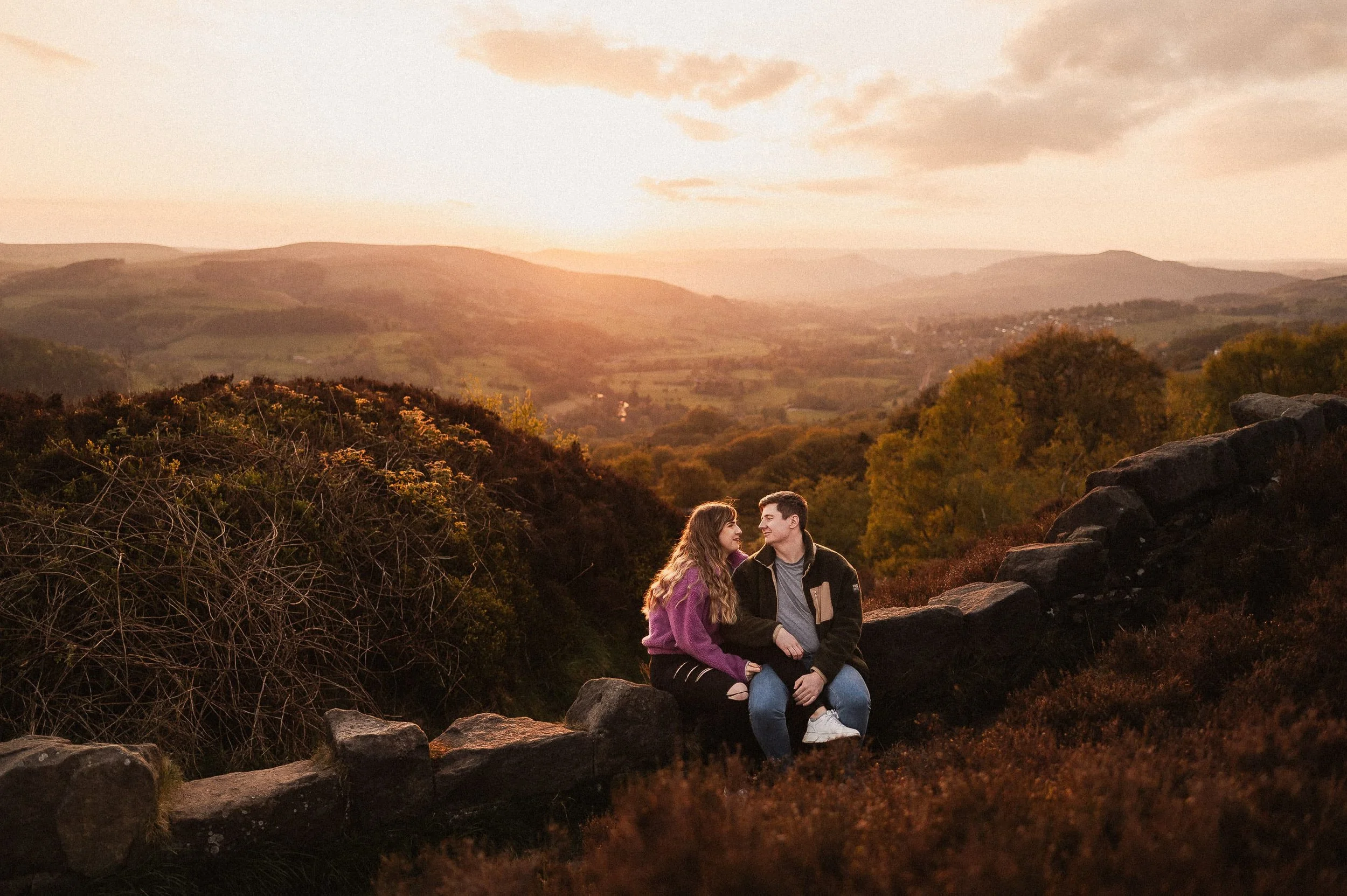 A young couple sitting on a stone wall in a scenic hilly landscape during sunset, looking at each other affectionately.