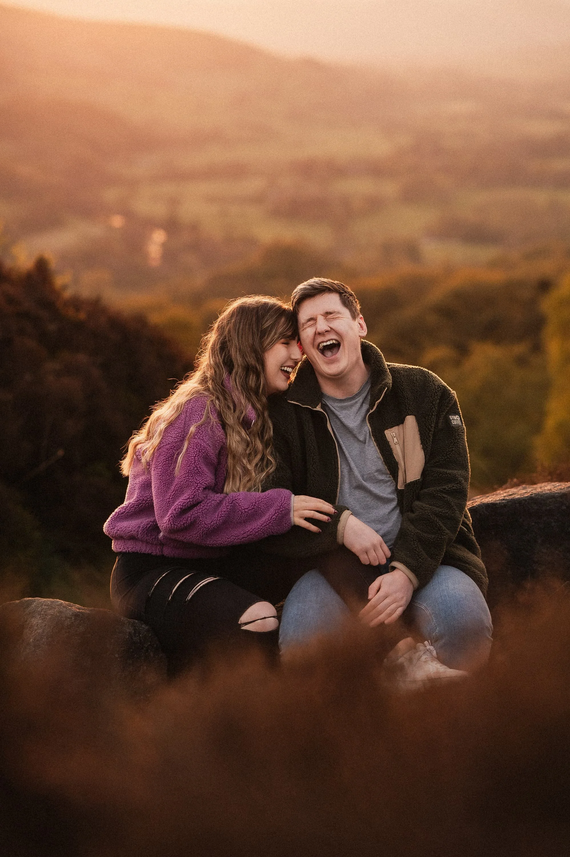 A young couple sitting outdoors on a rocky surface, laughing and enjoying each other's company during sunset with a blurred, scenic landscape in the background.