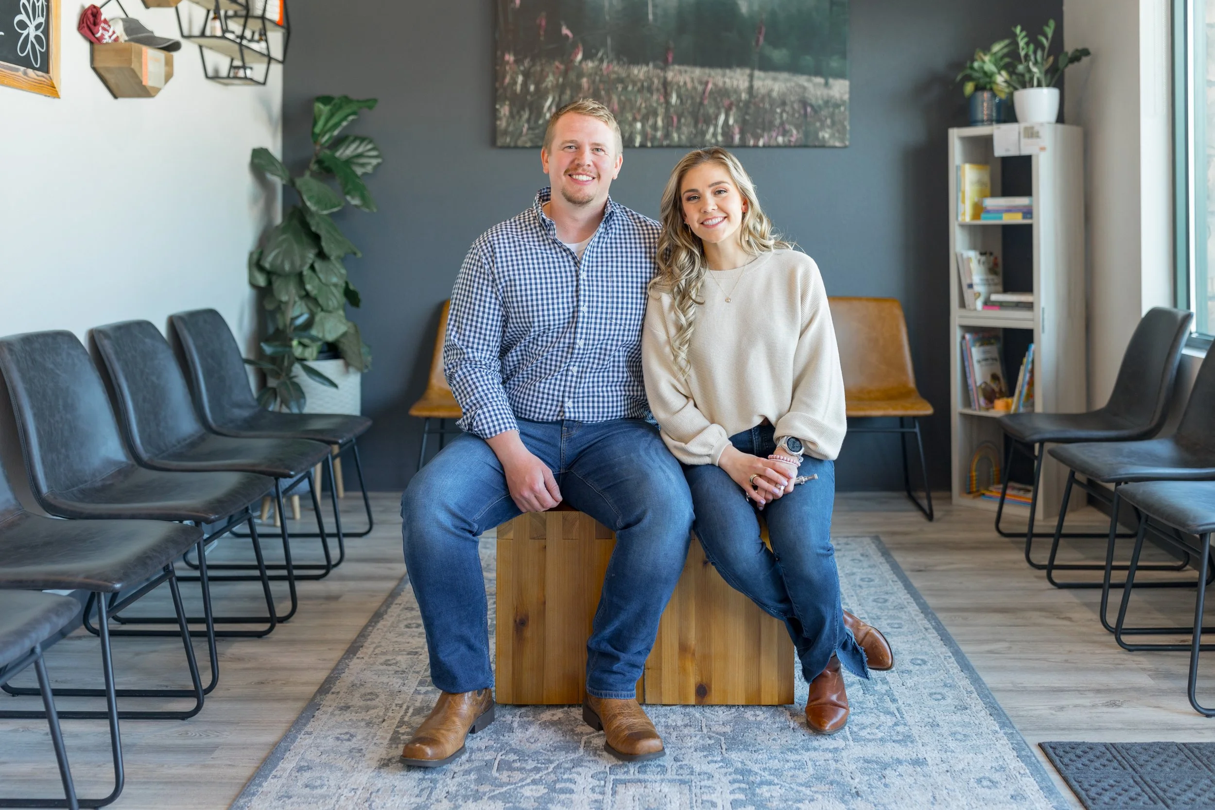 A man and woman smiling and sitting together on a wooden block in a waiting room, with black chairs and a bookshelf in the background.