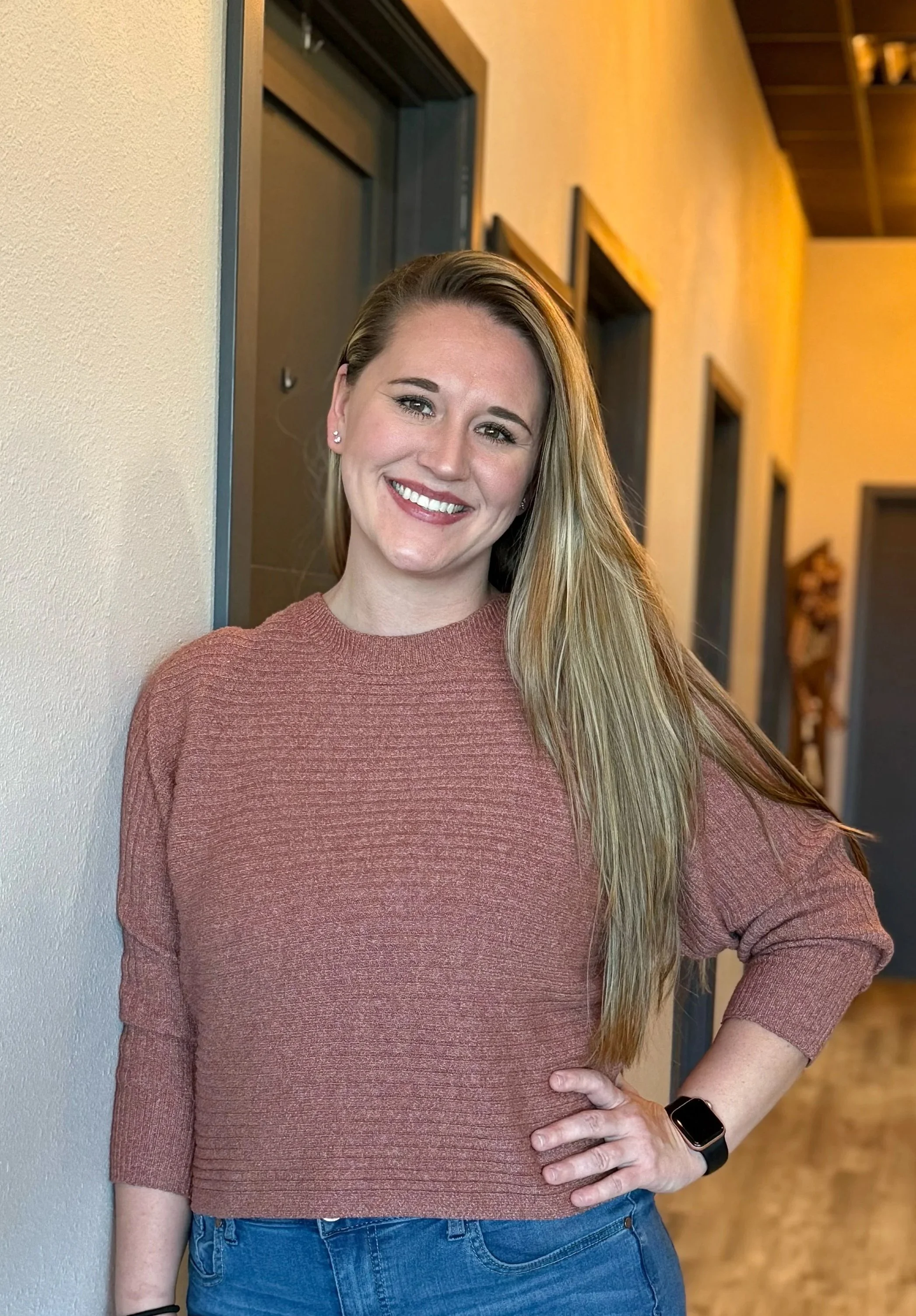 A woman with long blonde hair, wearing a pinkish-brown sweater, standing indoors against a beige wall, smiling with her hand on her hip.