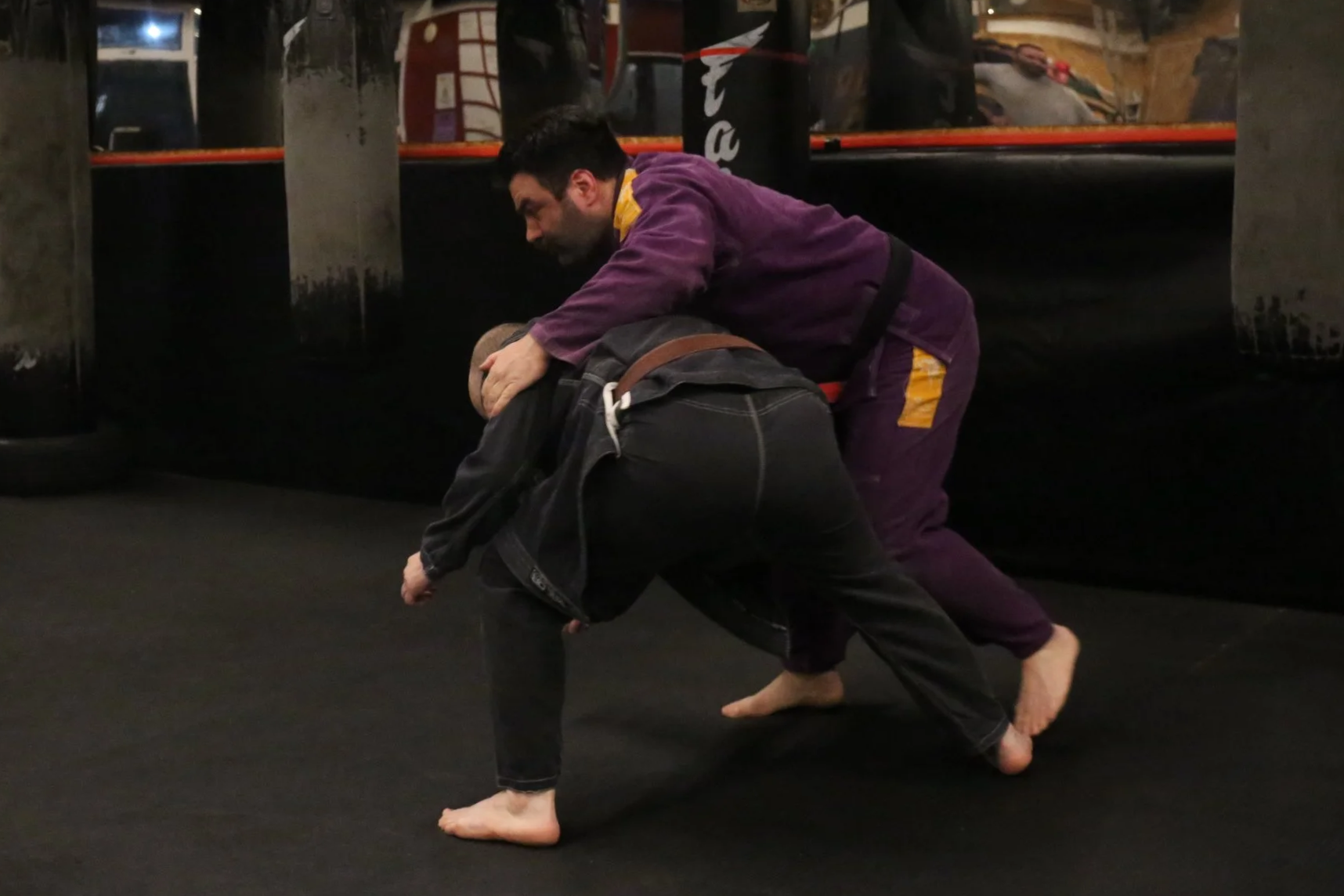 Two men practicing Brazilian Jiu-Jitsu grappling on a black mat, with one man in a purple gi and the other in black gi, engaging in aroused physical training in a gym.