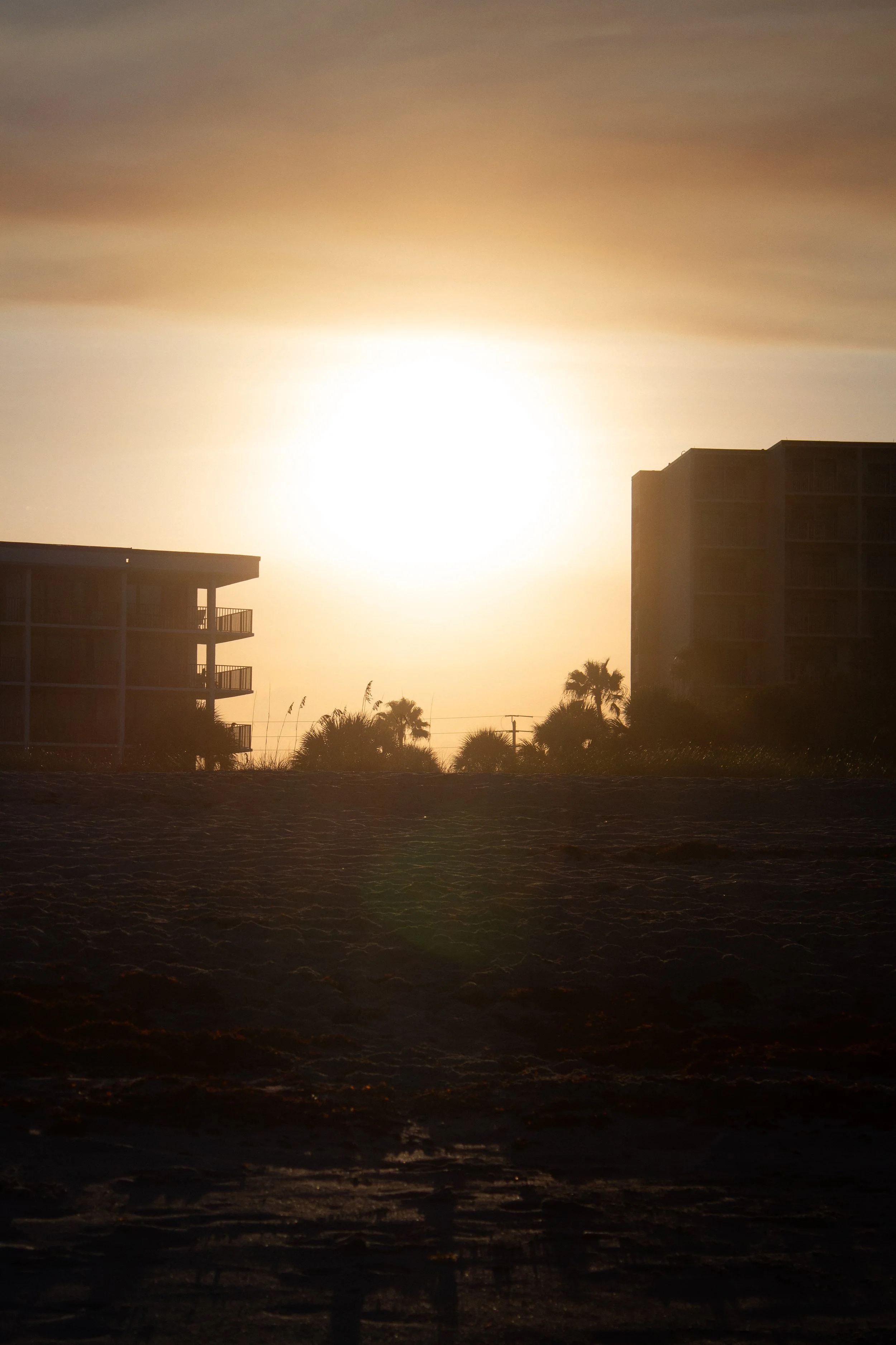 Sun setting over buildings and palm trees on a sandy beach.