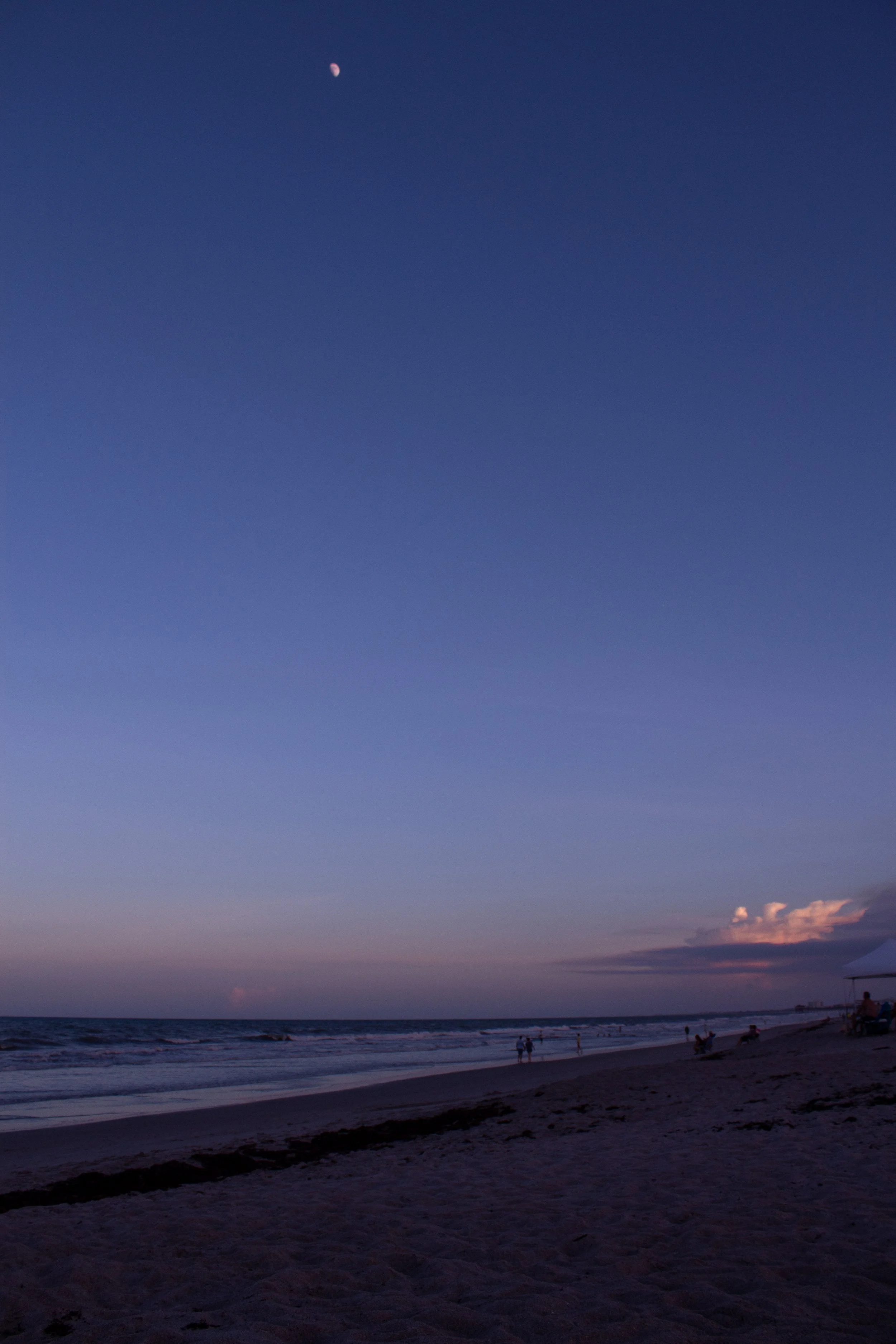 Beach scene at dusk with people walking and sitting along the shoreline, a tent on the right, and the moon in a clear sky with some clouds near the horizon.