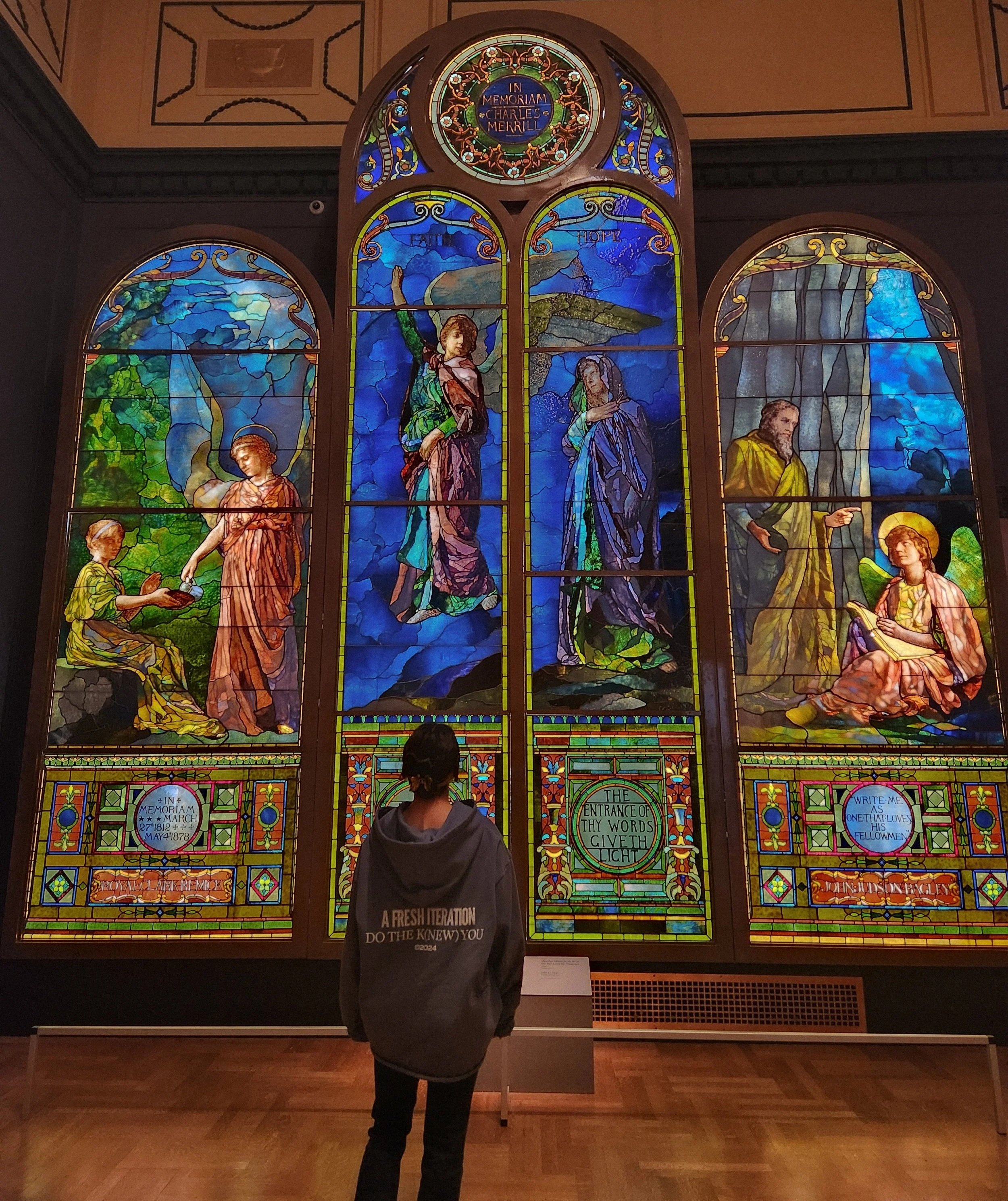 Person standing in front of a large colorful stained glass window depicting biblical scenes and figures.