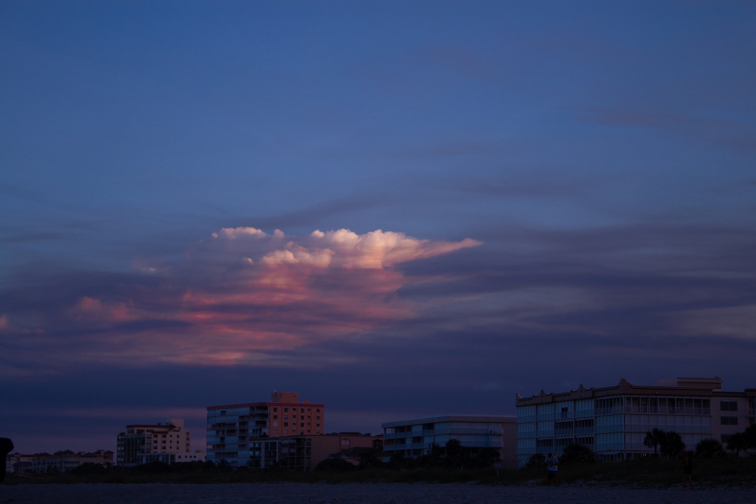 Sunset sky with pink and purple clouds over beachfront buildings and a few people walking.