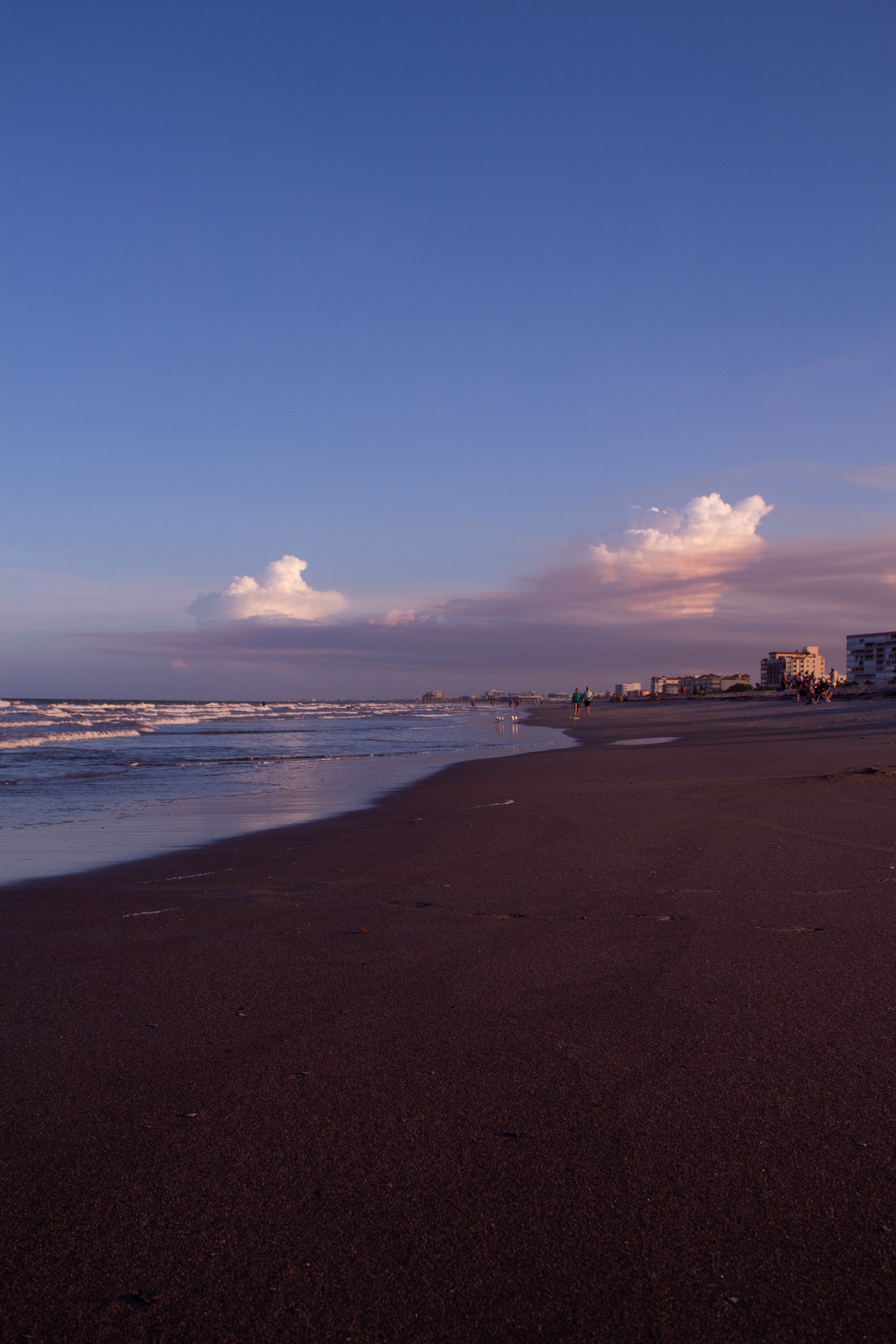 A sandy beach at sunset with a few people walking along the shoreline, some buildings in the distance, and a sky with clouds and a mix of blue and pink hues.