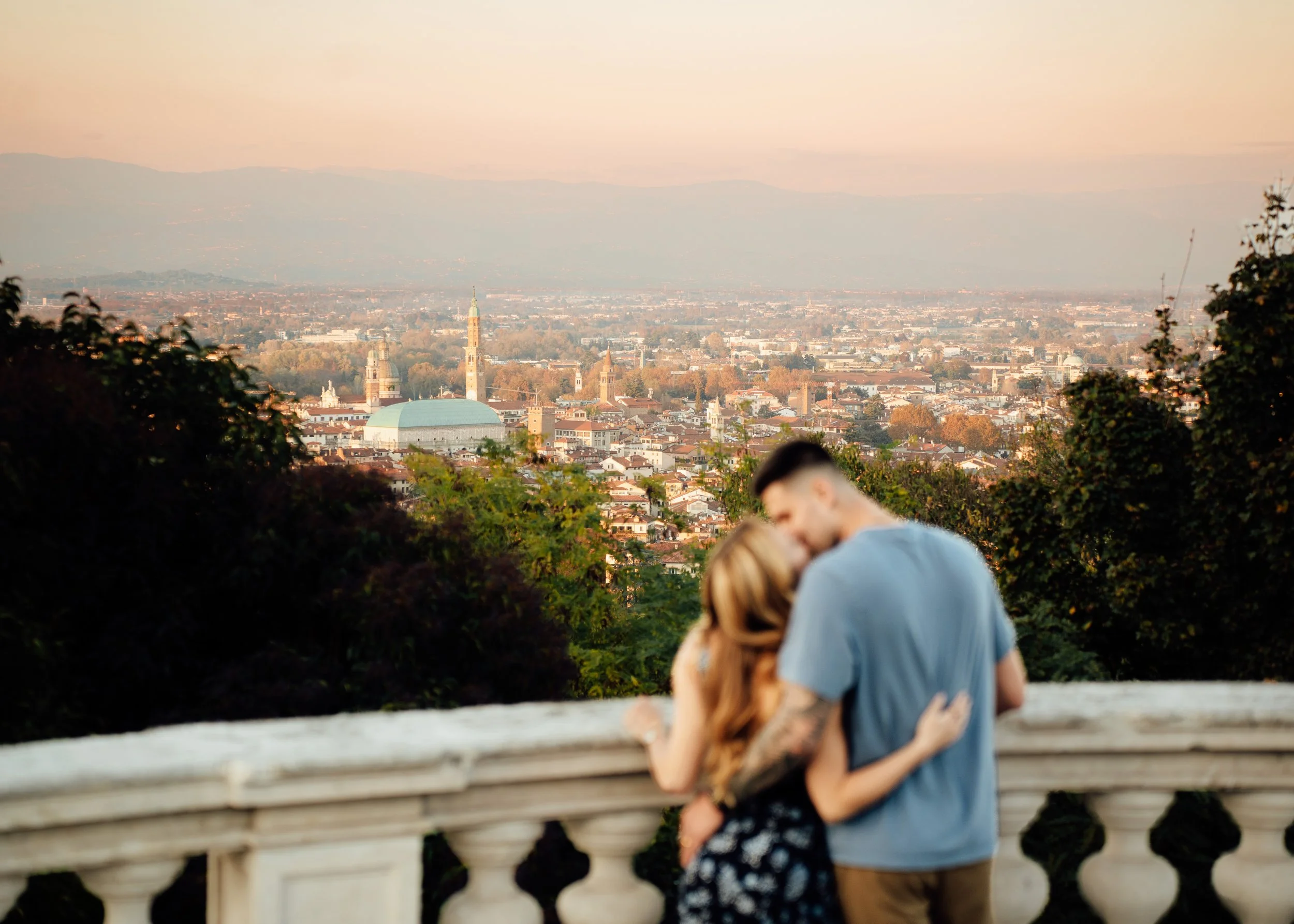 couple embracing and kissing at overlook with panoramic views of northern Italy skyline during sunset.
