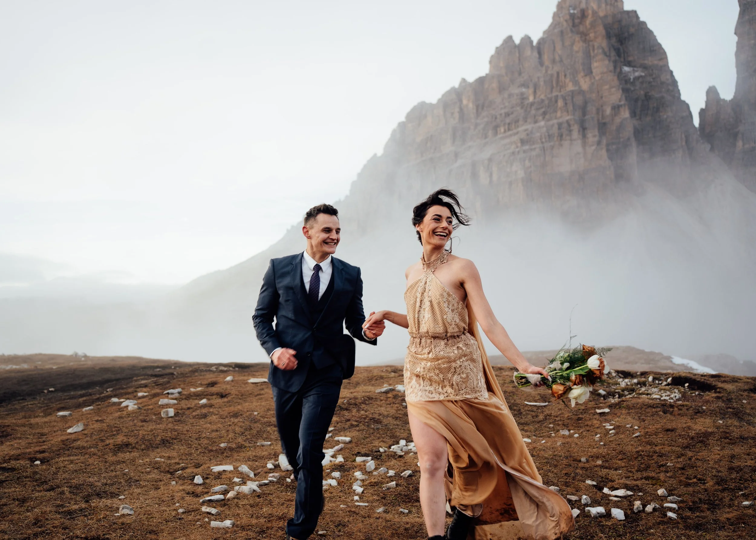 couple eloping at tre cime di lavaredo holding hands running in wedding clothes and bouquet of flowers